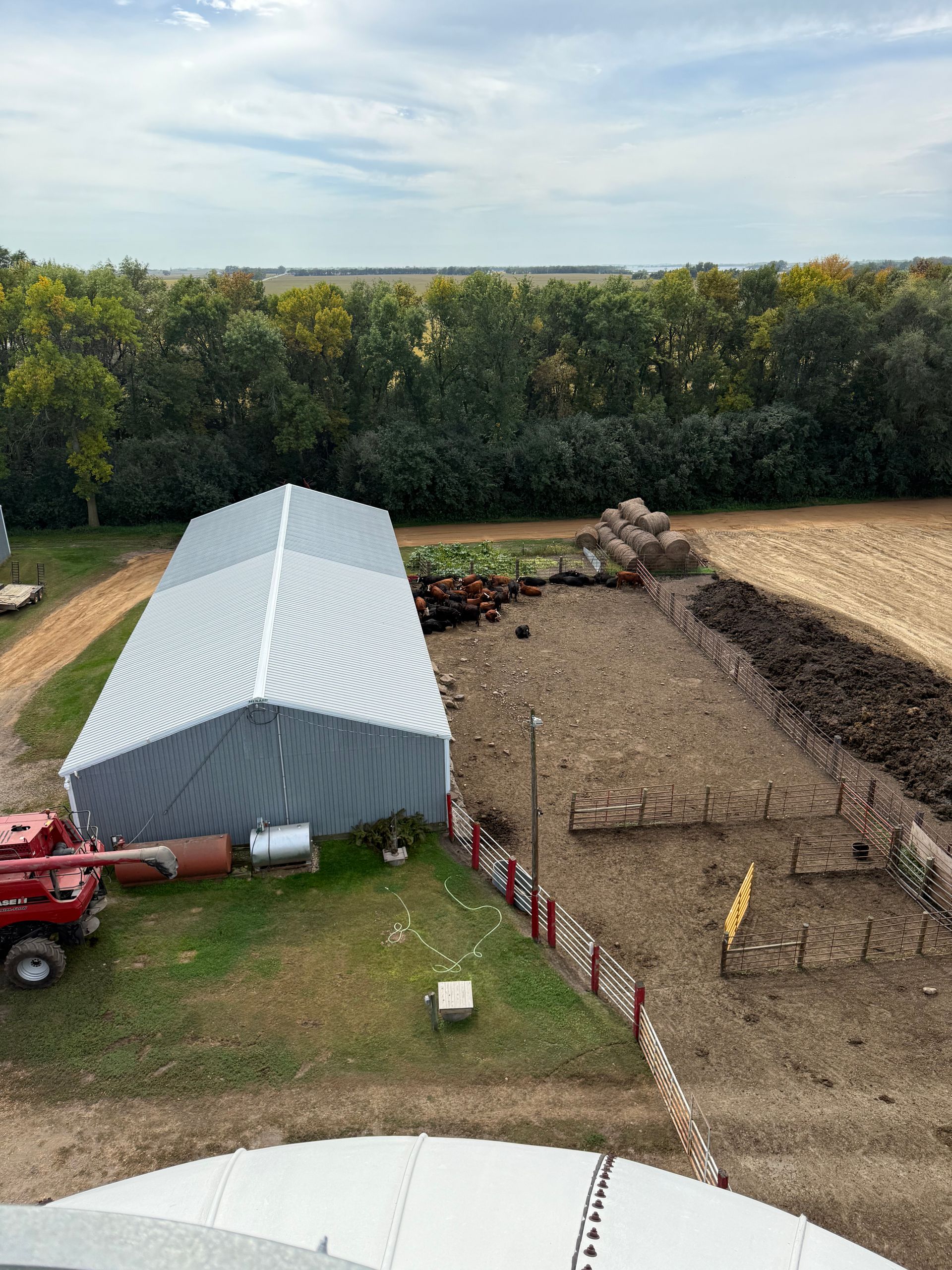Farm scene: A metal-roofed building, cattle pen, and tractor near trees and field under a cloudy sky.