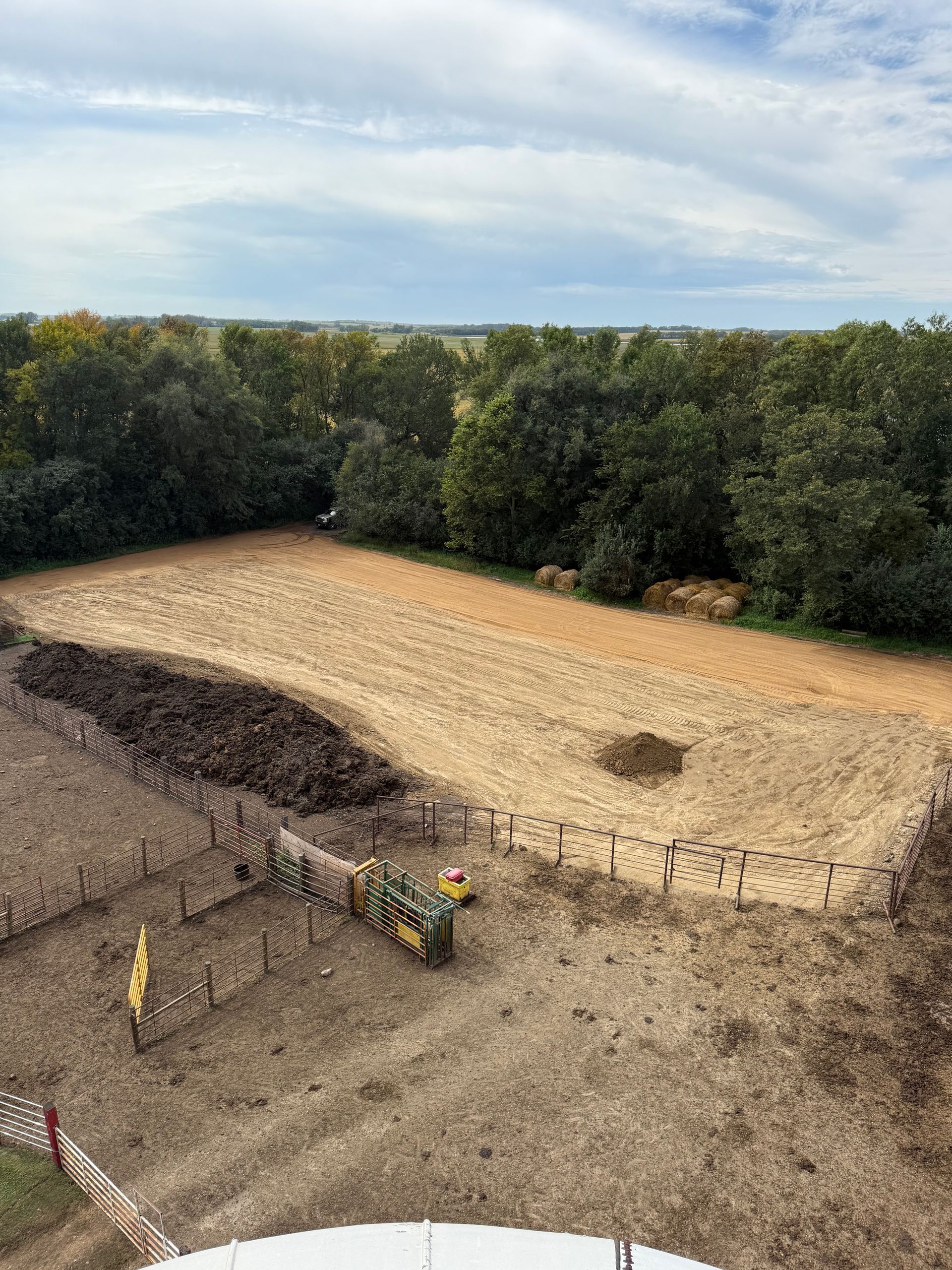Aerial view of a field, partially tilled, with a tractor and large pile of dark material in the foreground, trees in the background.