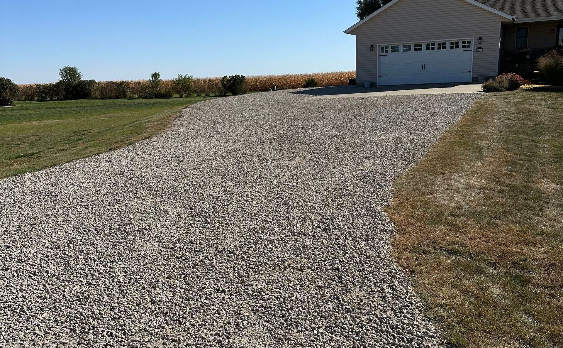 Gravel driveway leading to a garage next to green grass and a field on a sunny day.