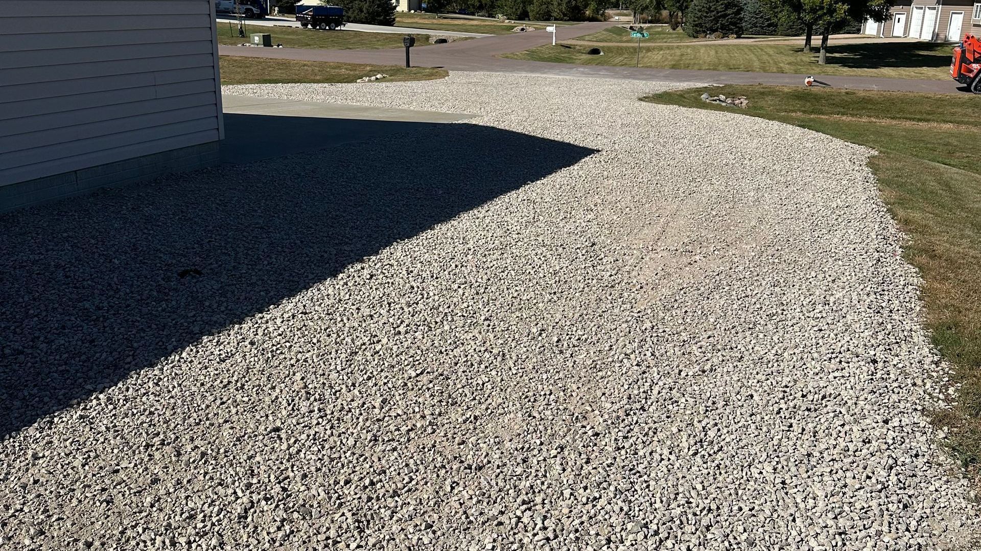Gravel driveway next to a building, partially in shadow, leading to a street and grassy area.