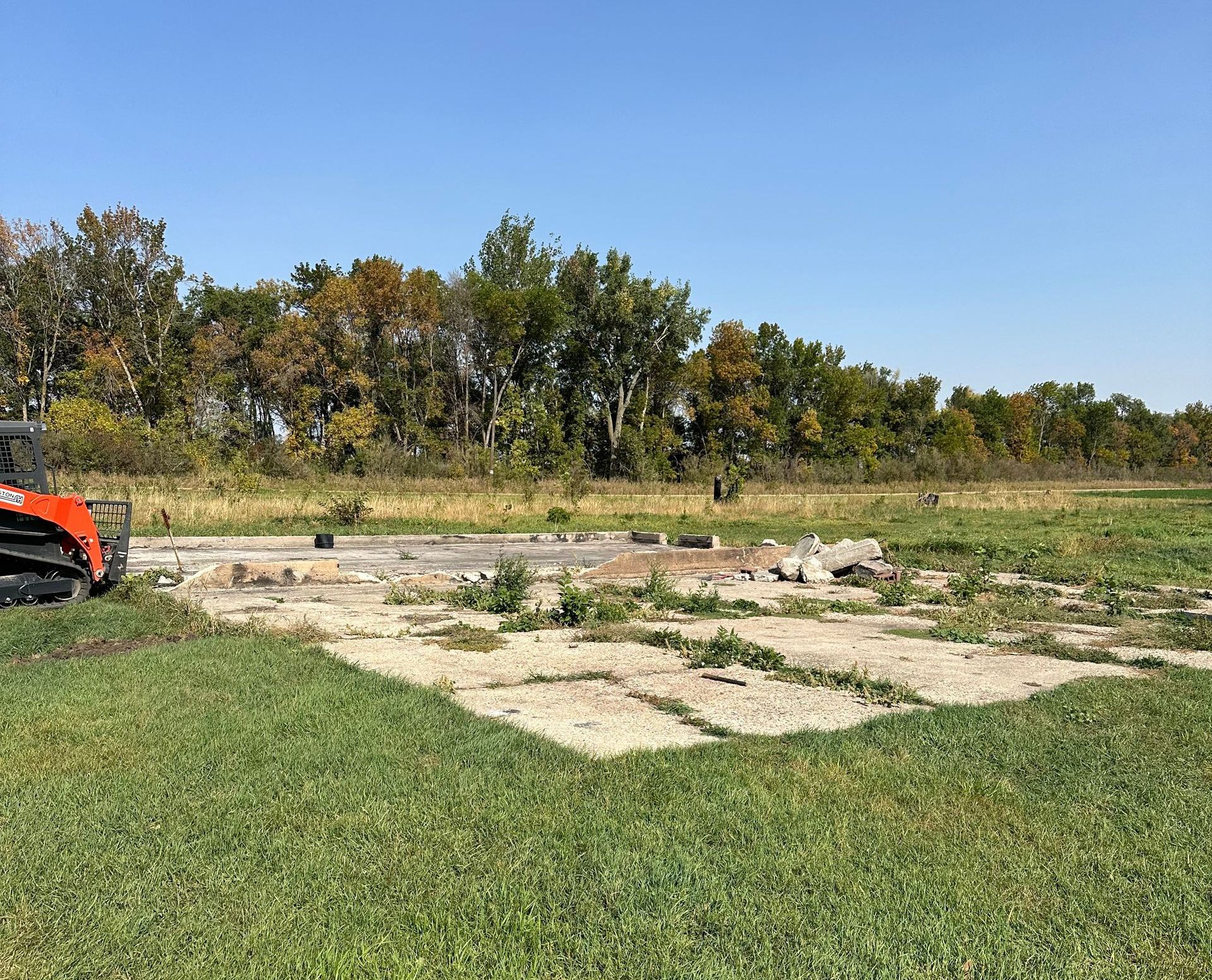 Remains of foundation in a grassy field with trees under a blue sky; an orange skid steer sits at the left.