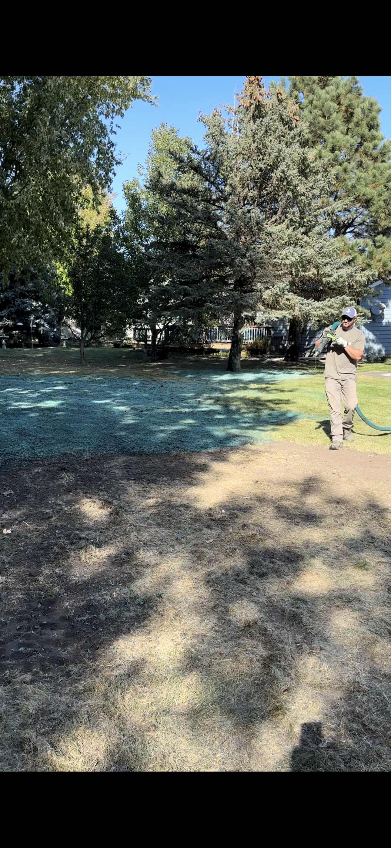A person in beige clothes walks in a grassy yard towards the camera; trees are behind them.