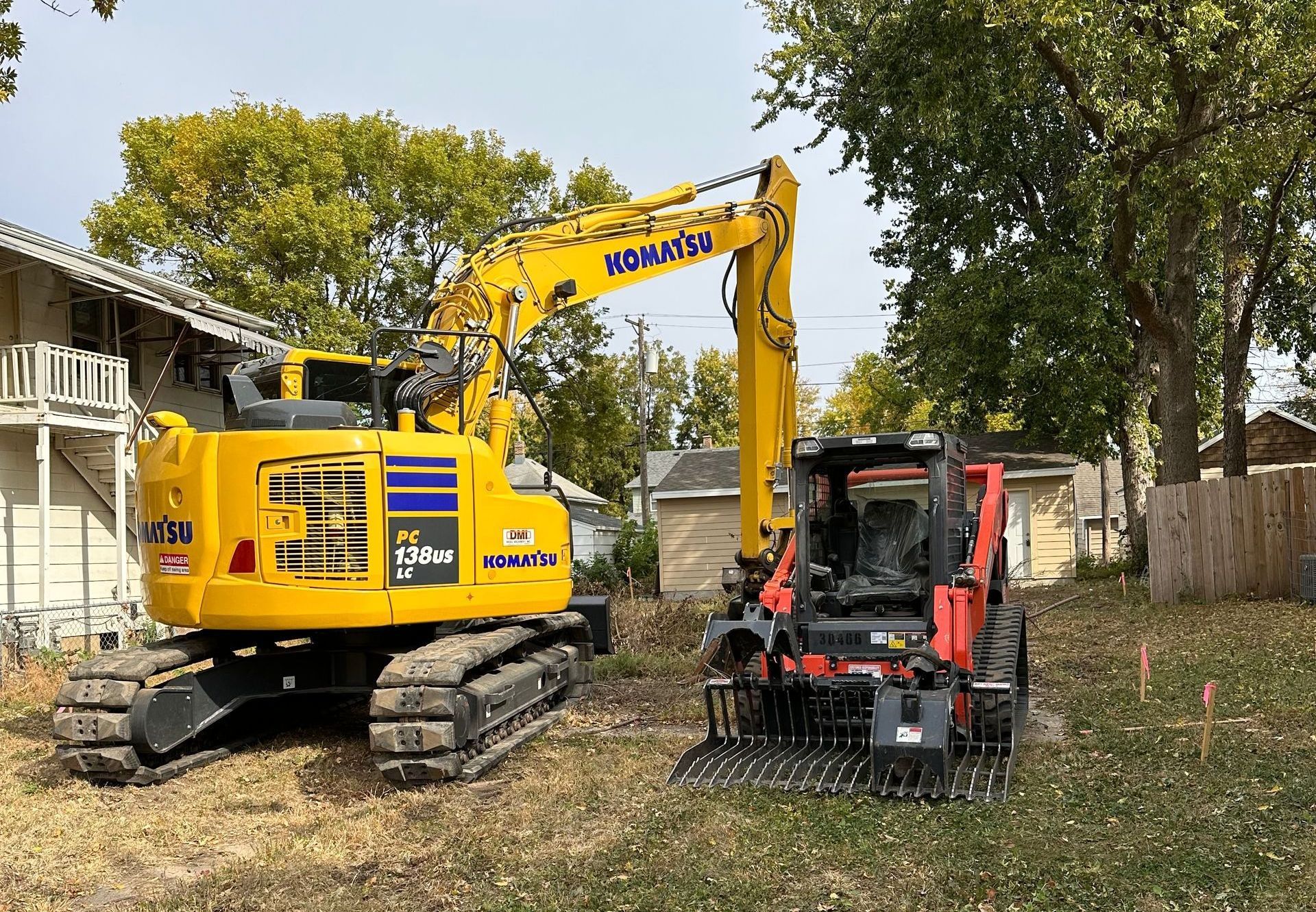 Yellow Komatsu excavator and orange Kubota skid steer on a grassy lot, near a building.