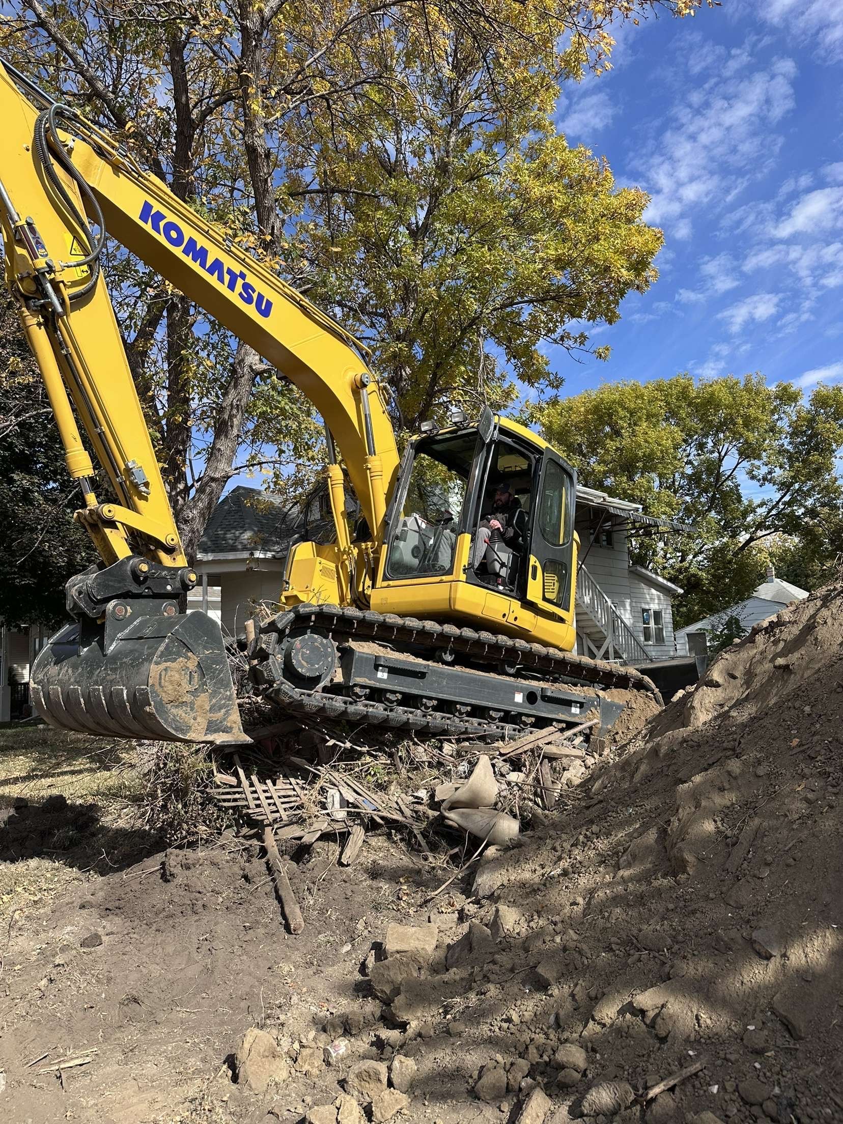Yellow Komatsu excavator digging earth in a yard, trees in background, sunny day.