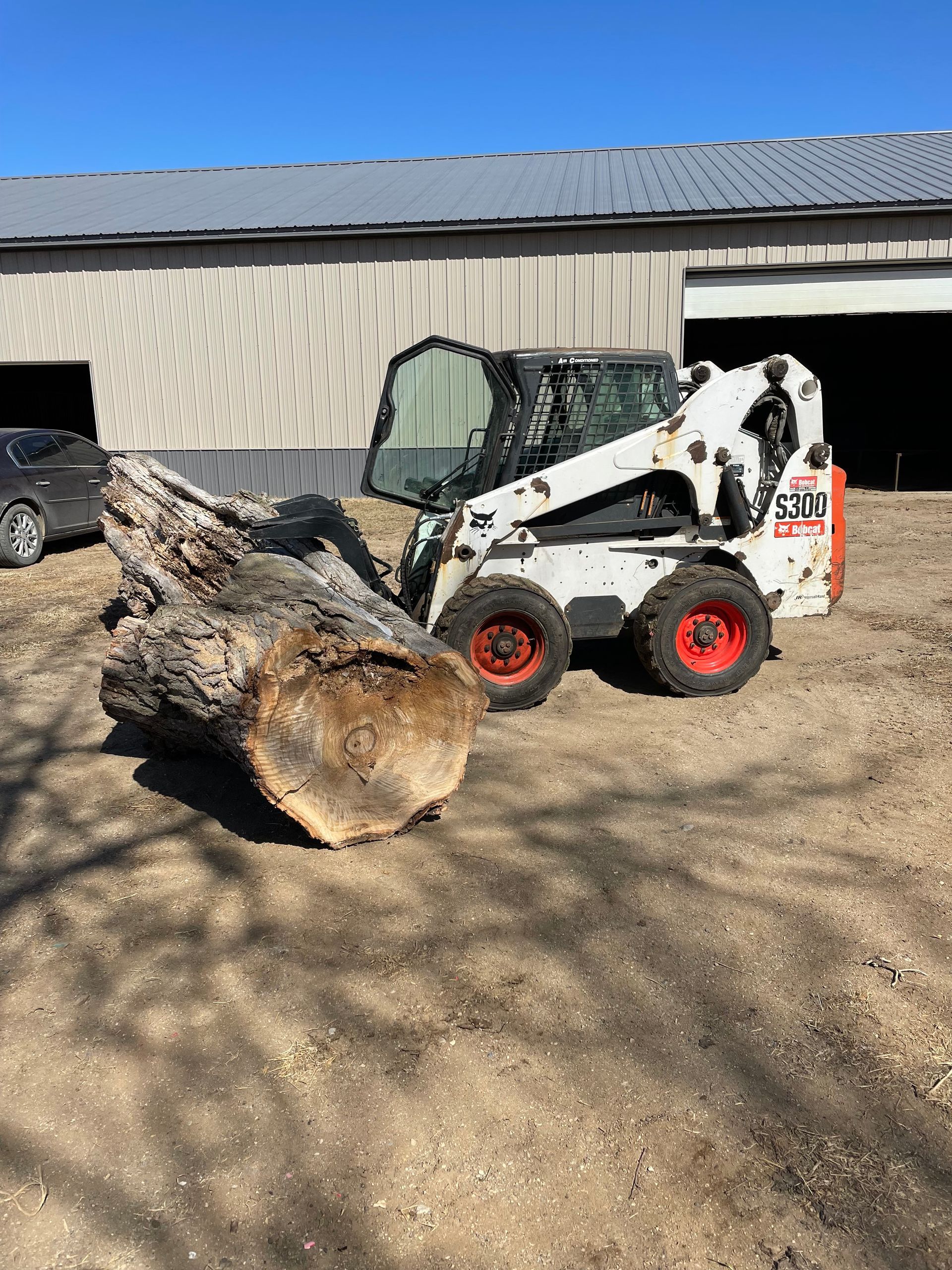 Bobcat skid steer with a large log outdoors near a barn.