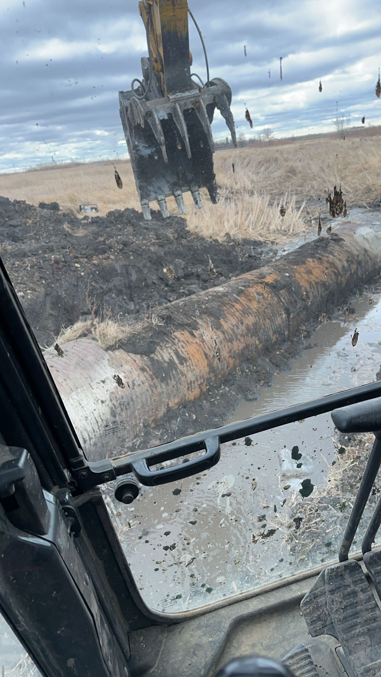 An excavator digs in muddy ground, revealing a large pipe. View is from the operator's cab.