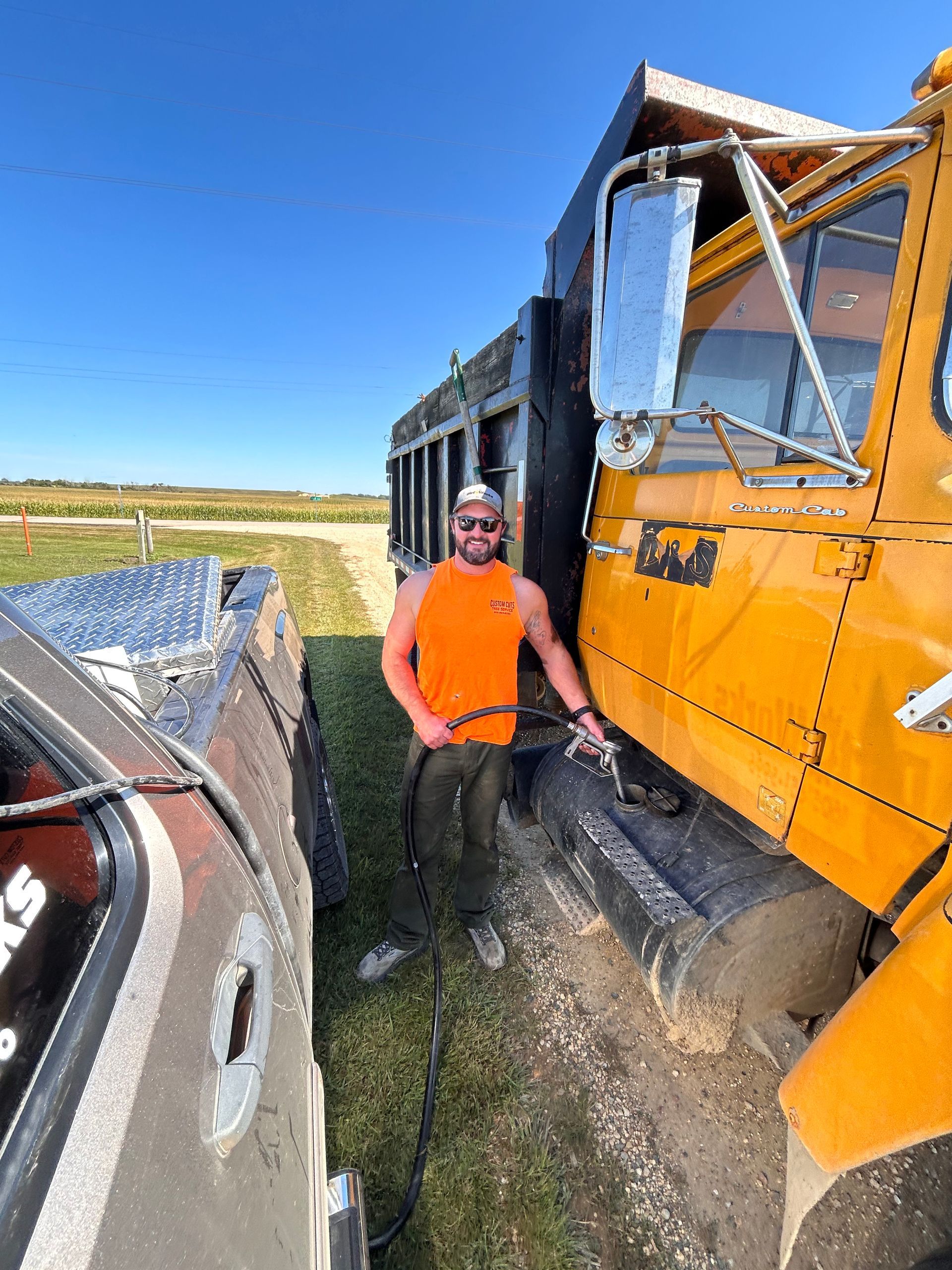 Man in orange shirt next to yellow dump truck, towing it with a silver truck in a field.