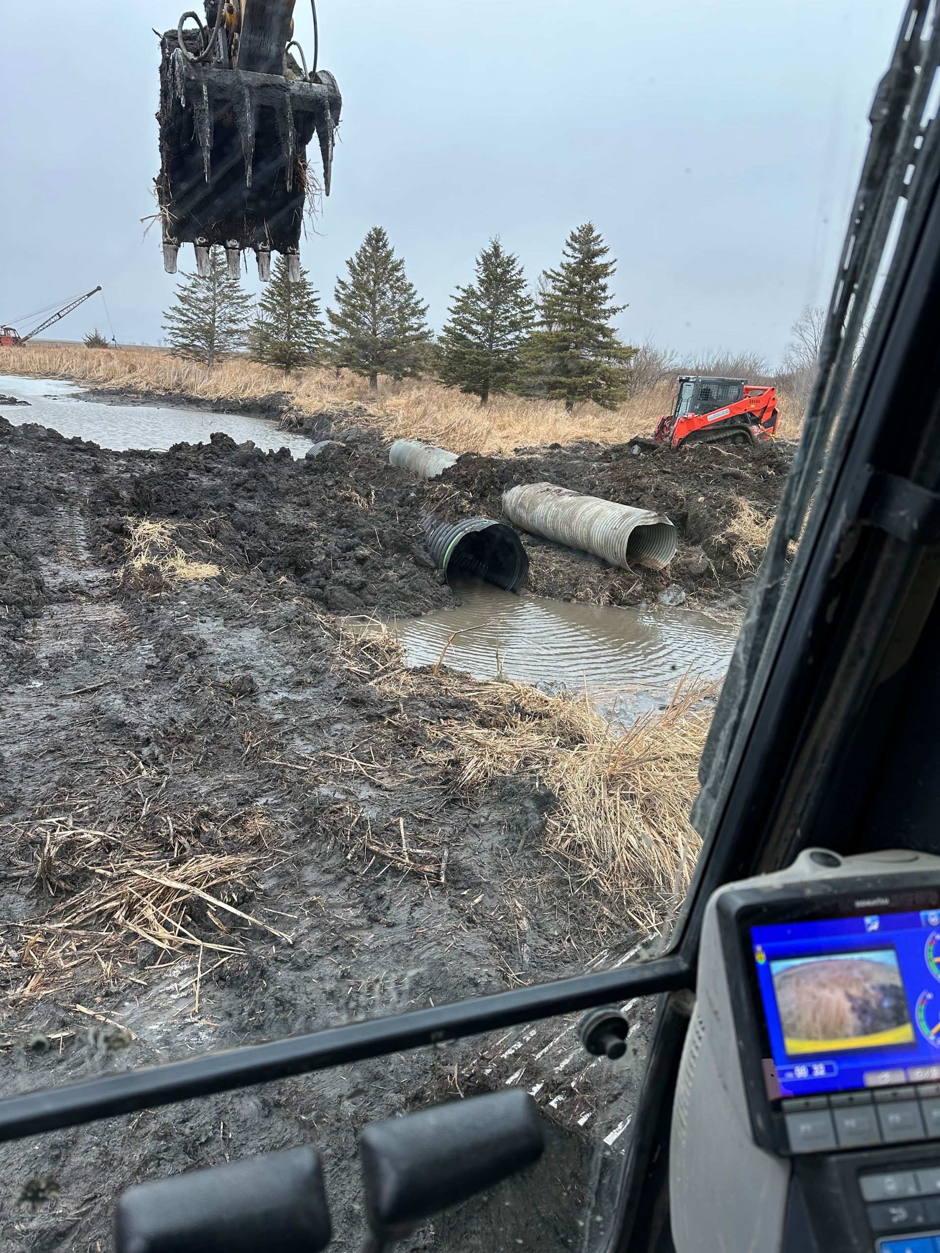 An excavator placing a concrete pipe in a muddy construction site, with another machine in the distance.