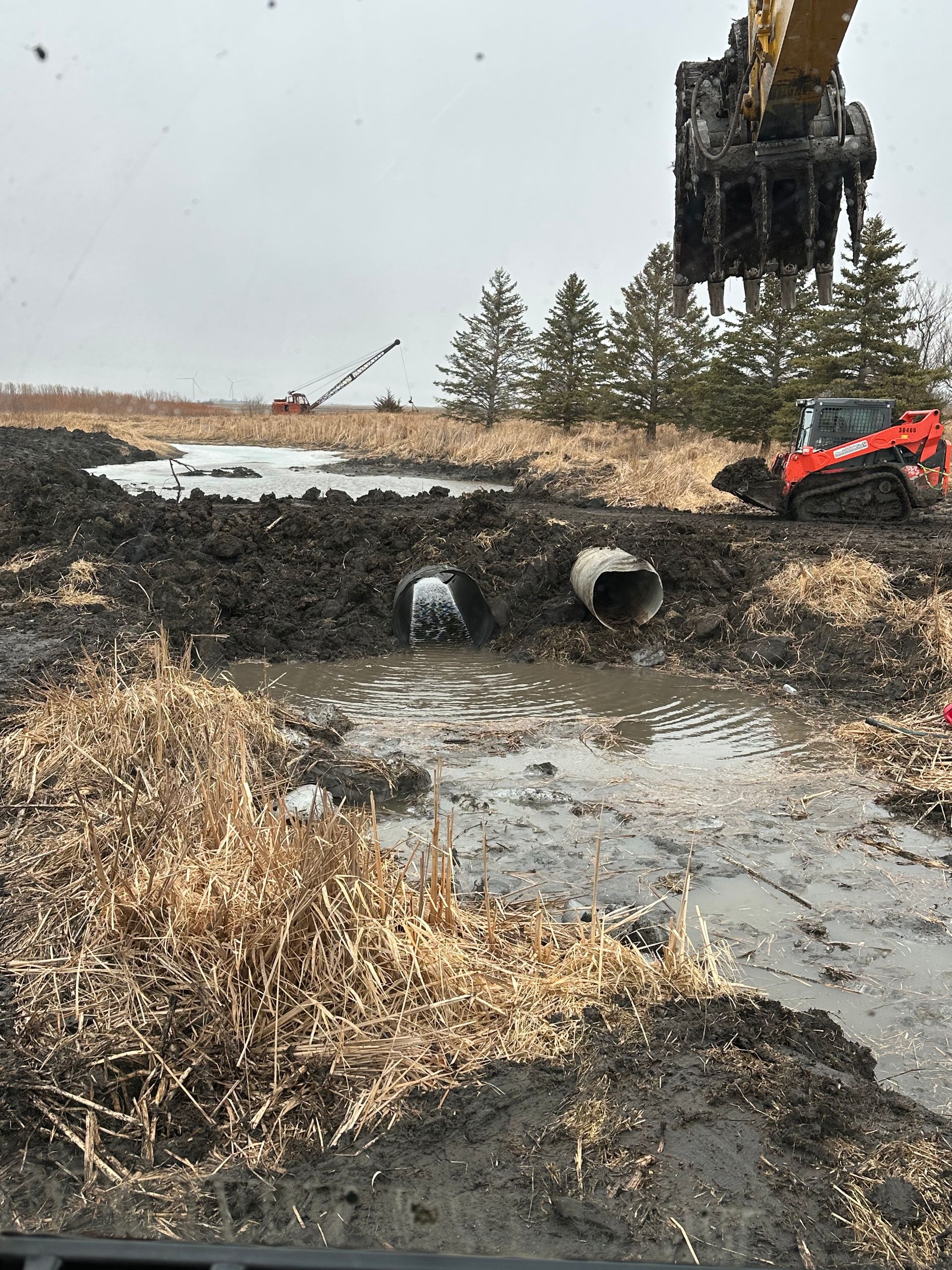 Construction site with excavator removing soil, pipes in muddy water. Red skid steer in background.