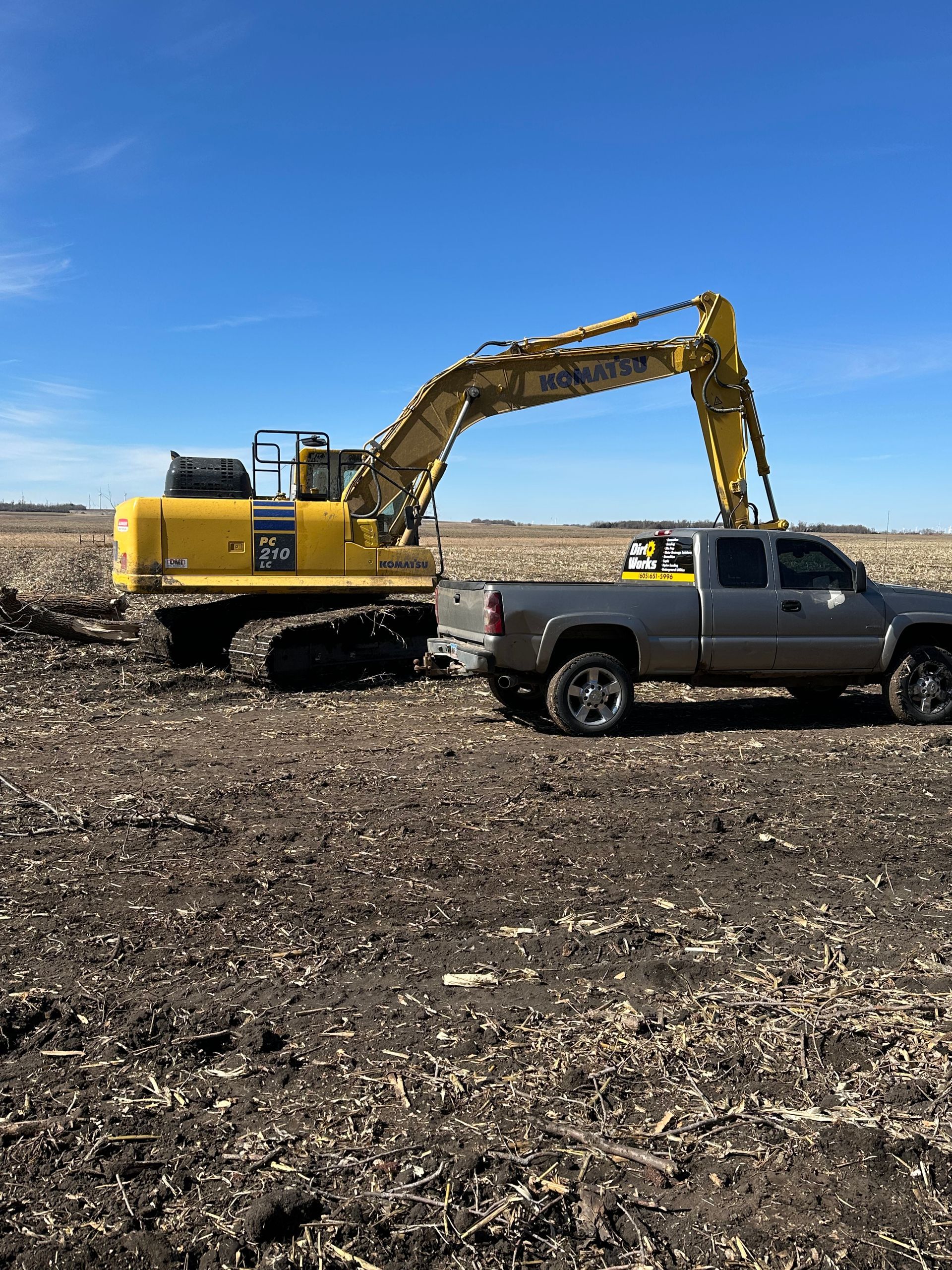 Yellow excavator and gray pickup truck in a field under a blue sky.