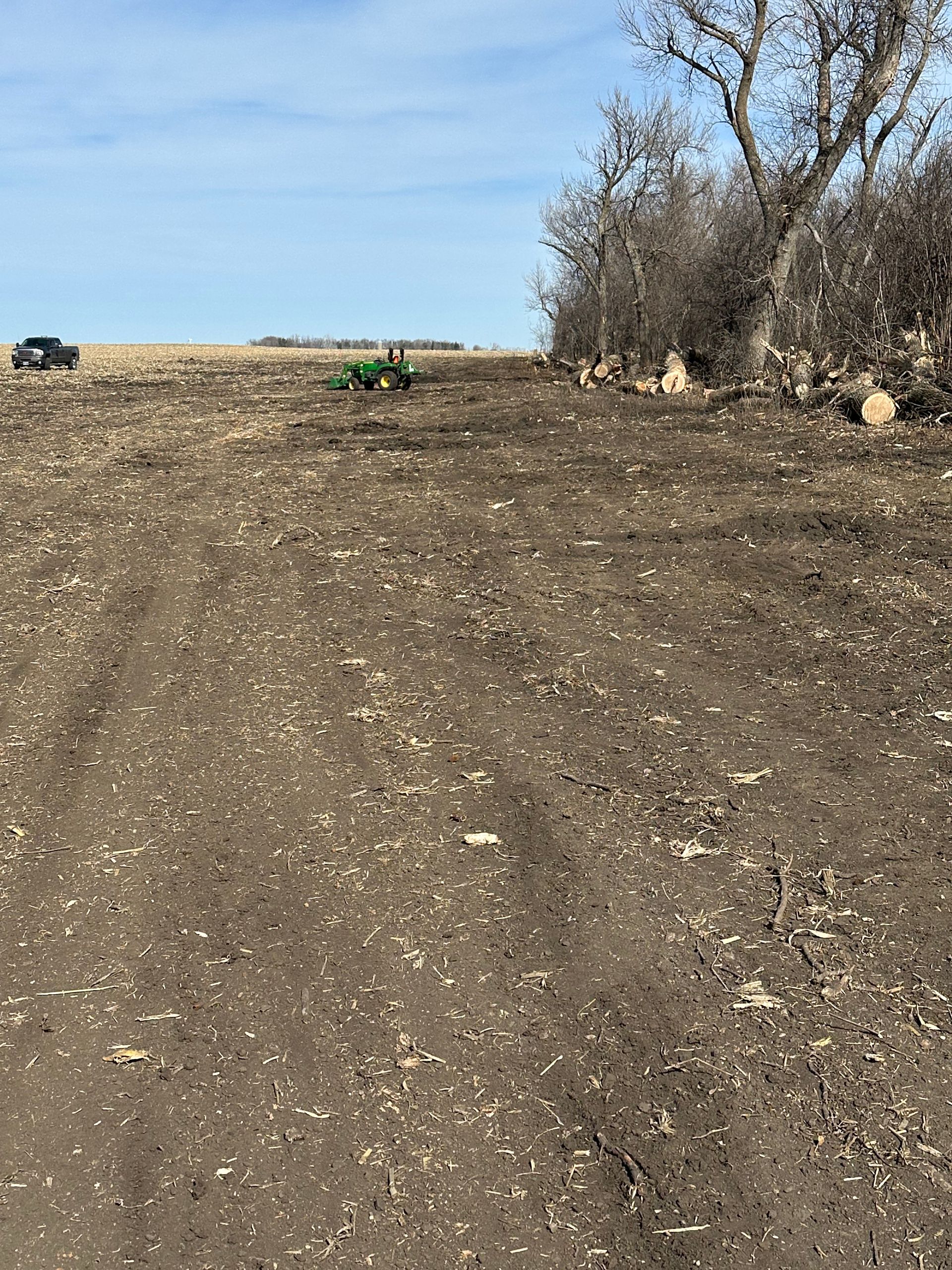 Dirt field with a tractor in the distance, blue sky, and a line of bare trees on the right.