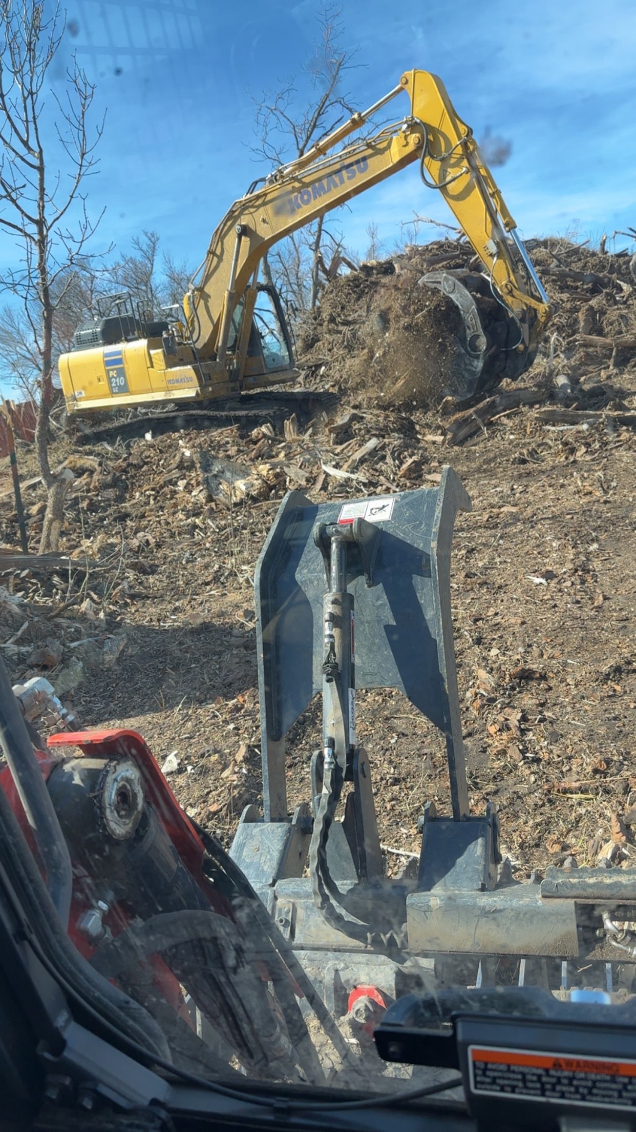A yellow excavator scoops debris on a hillside, viewed from inside a vehicle with a black attachment.