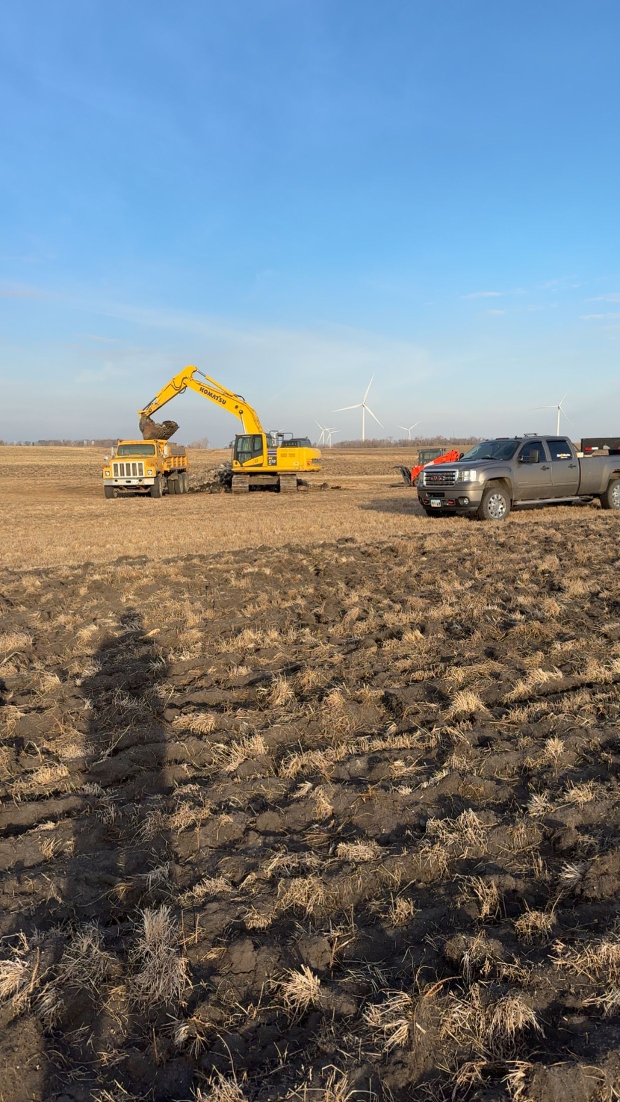 Excavator loading dirt into a dump truck in a field, with a pickup truck nearby and wind turbines in the distance.