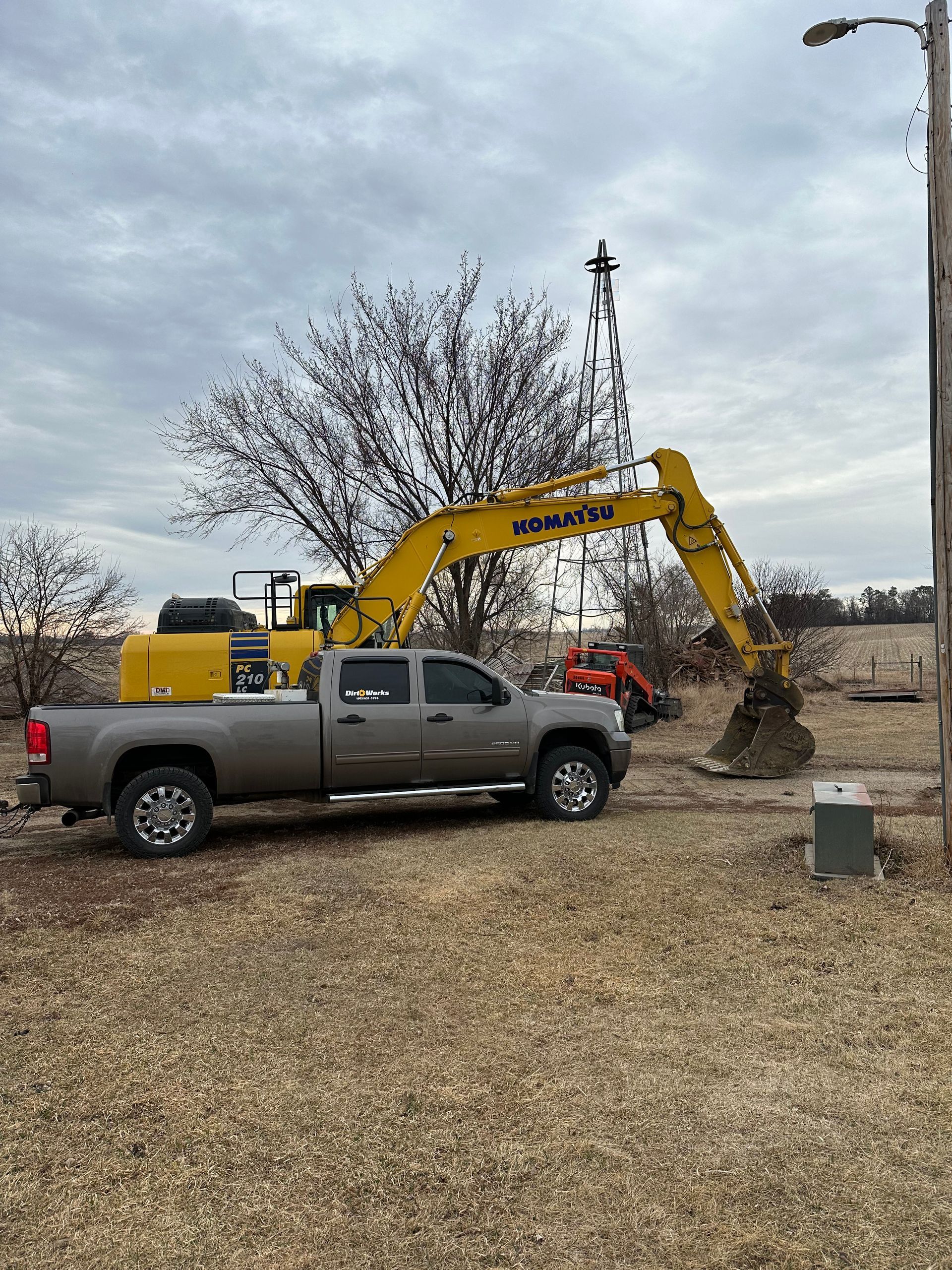 A gray pickup truck parked near a yellow excavator digging in a field under a cloudy sky.