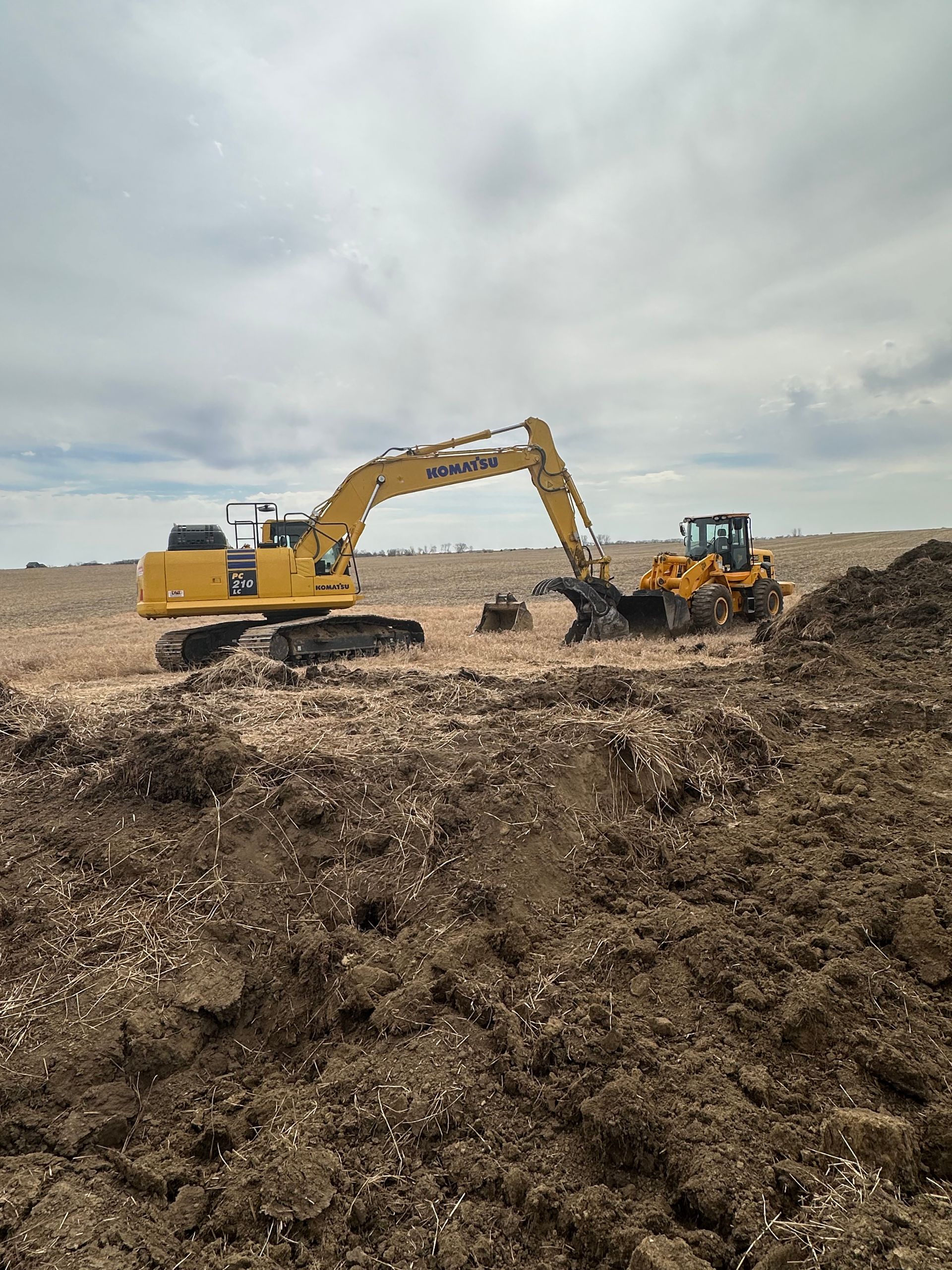 Excavator and loader on a construction site; overcast sky. The excavator digs while the loader waits nearby.