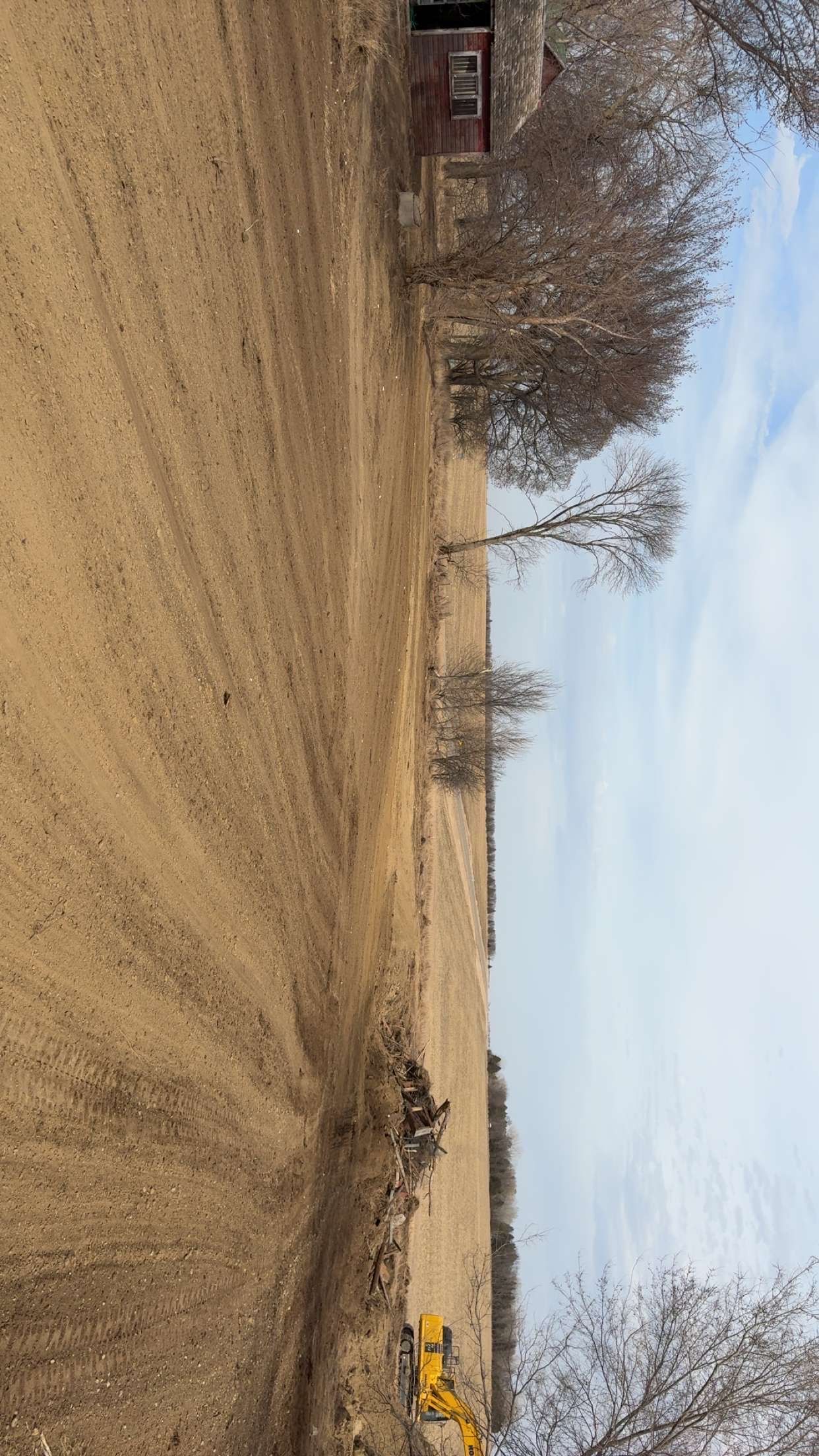 Brown field with a small building in the distance and bare trees under a cloudy sky.