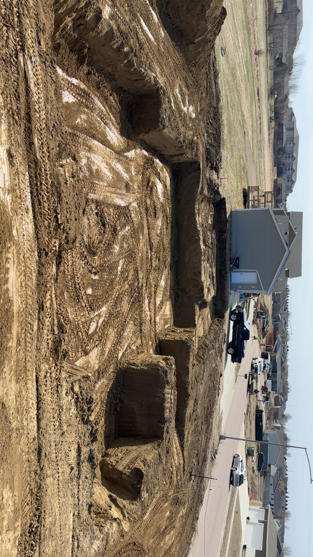Construction site with trenches dug for a foundation, near an existing house and road.