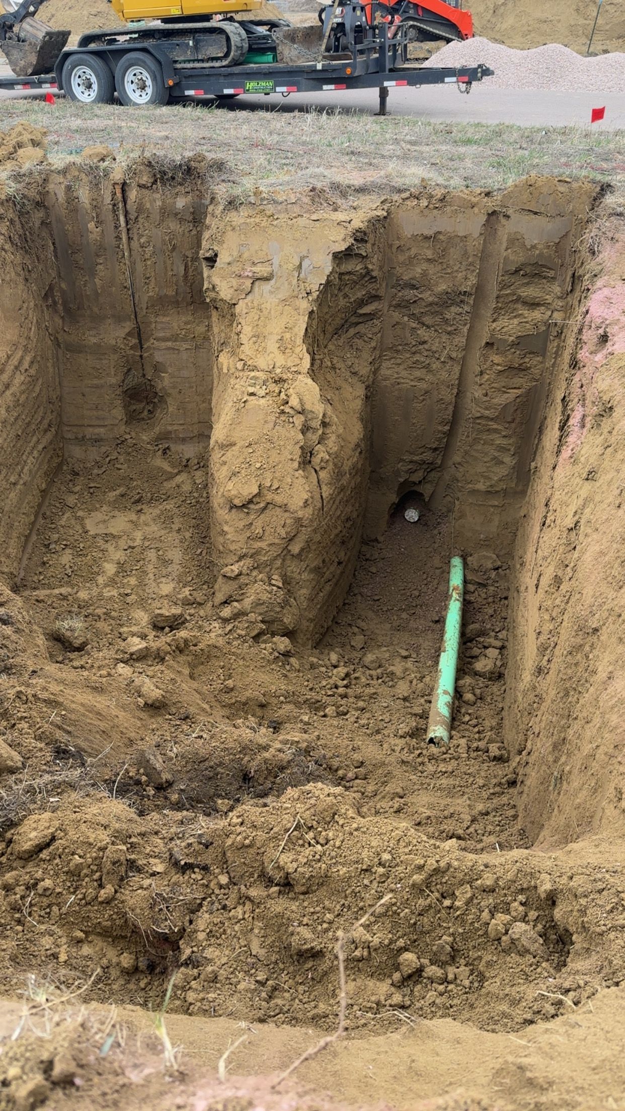 Excavated trench in dirt with visible green pipe; machinery and trailer in background.