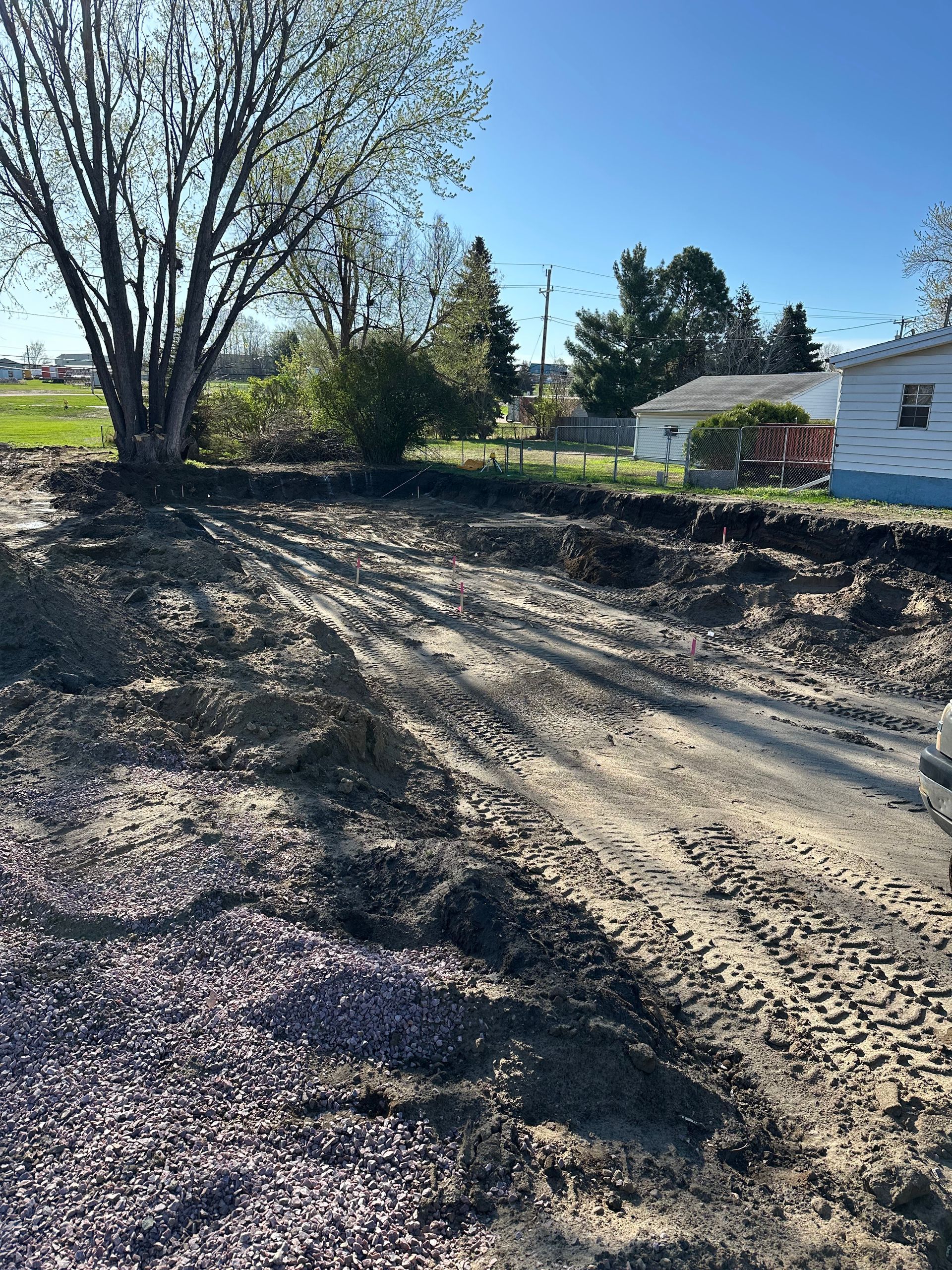 Dirt and gravel lot with tire tracks, tree, and house on a sunny day.