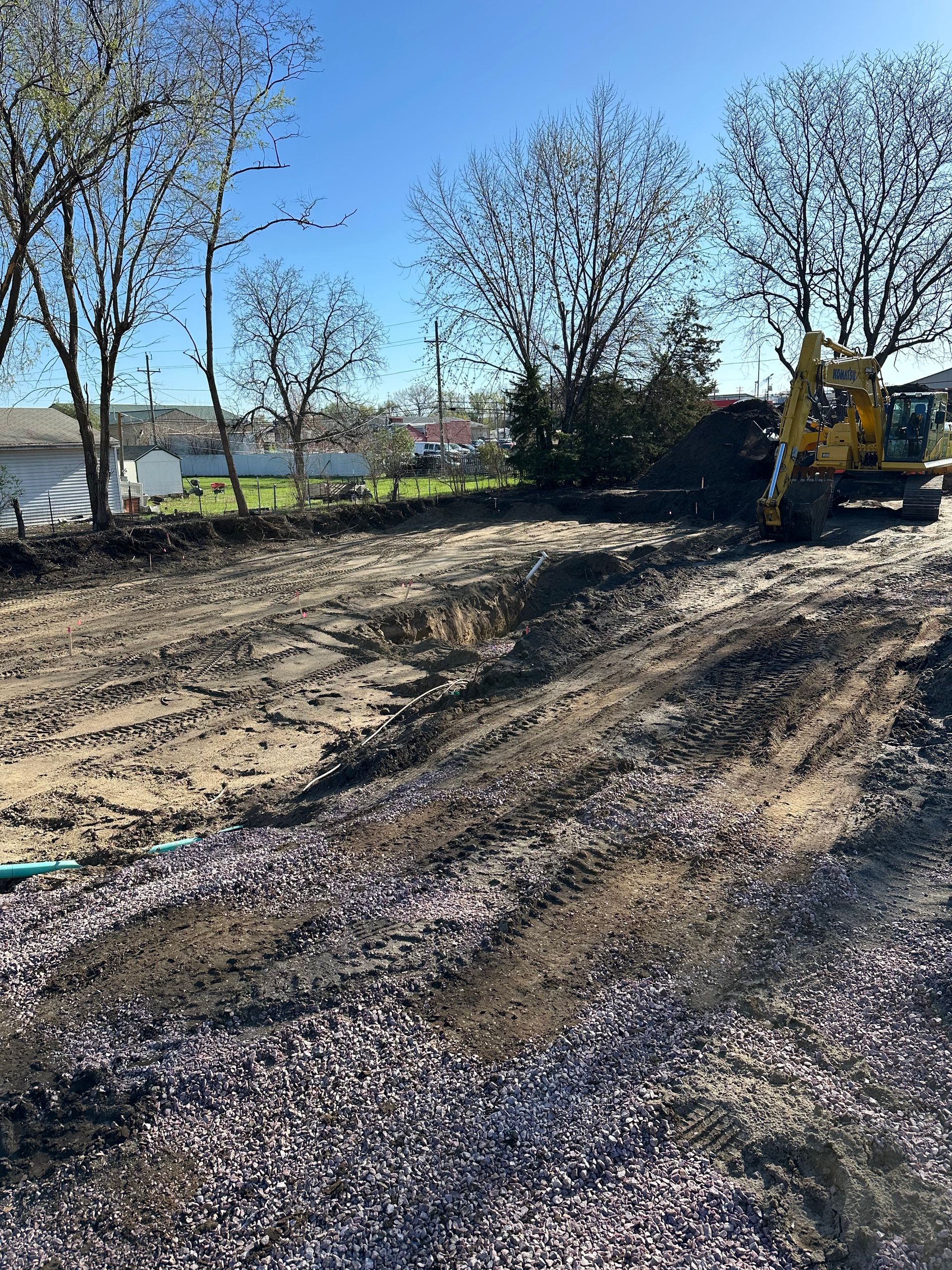 Construction site with an excavator, freshly turned earth, and gravel. Bare trees in the background.
