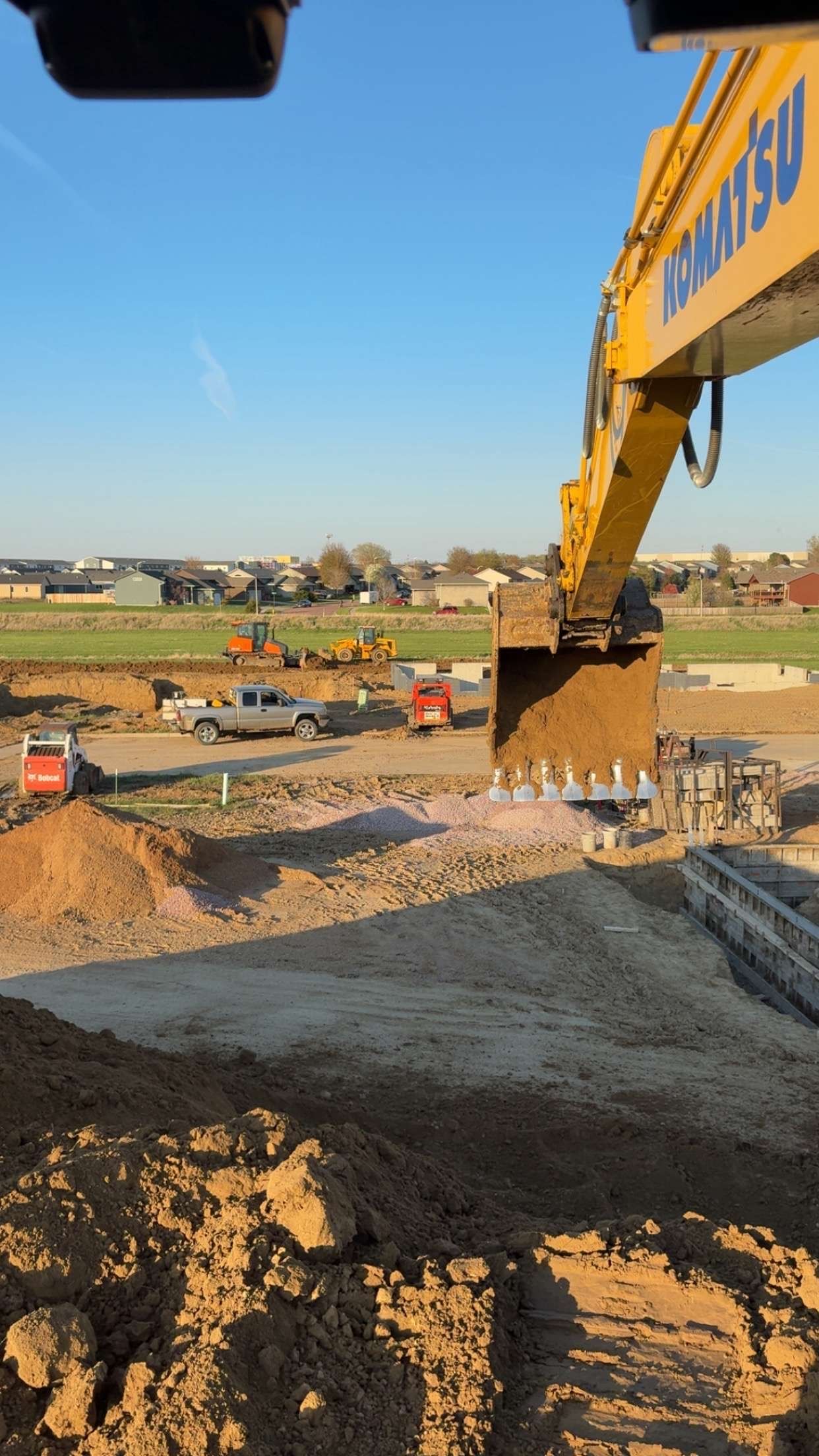 A Komatsu excavator at a construction site scoops up soil, other vehicles and structures in the background, blue sky.
