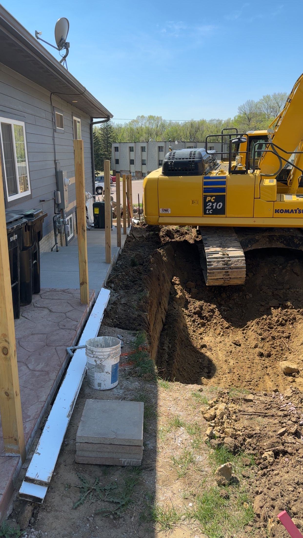 Yellow excavator digging a trench next to a building.  Dirt pile and construction materials are visible. Sunny day.