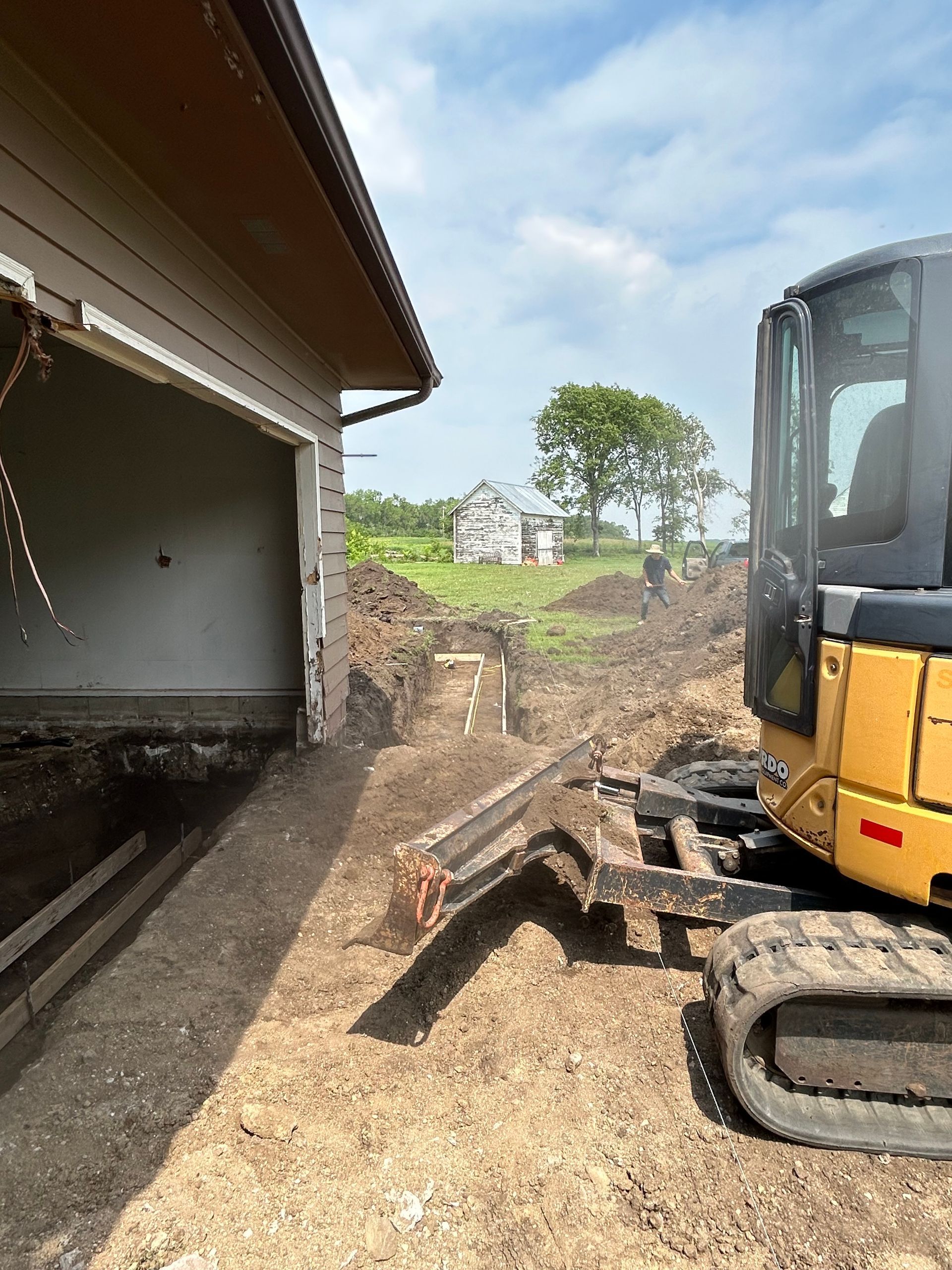 A small excavator digs near a garage, creating a trench in the dirt under a blue sky.