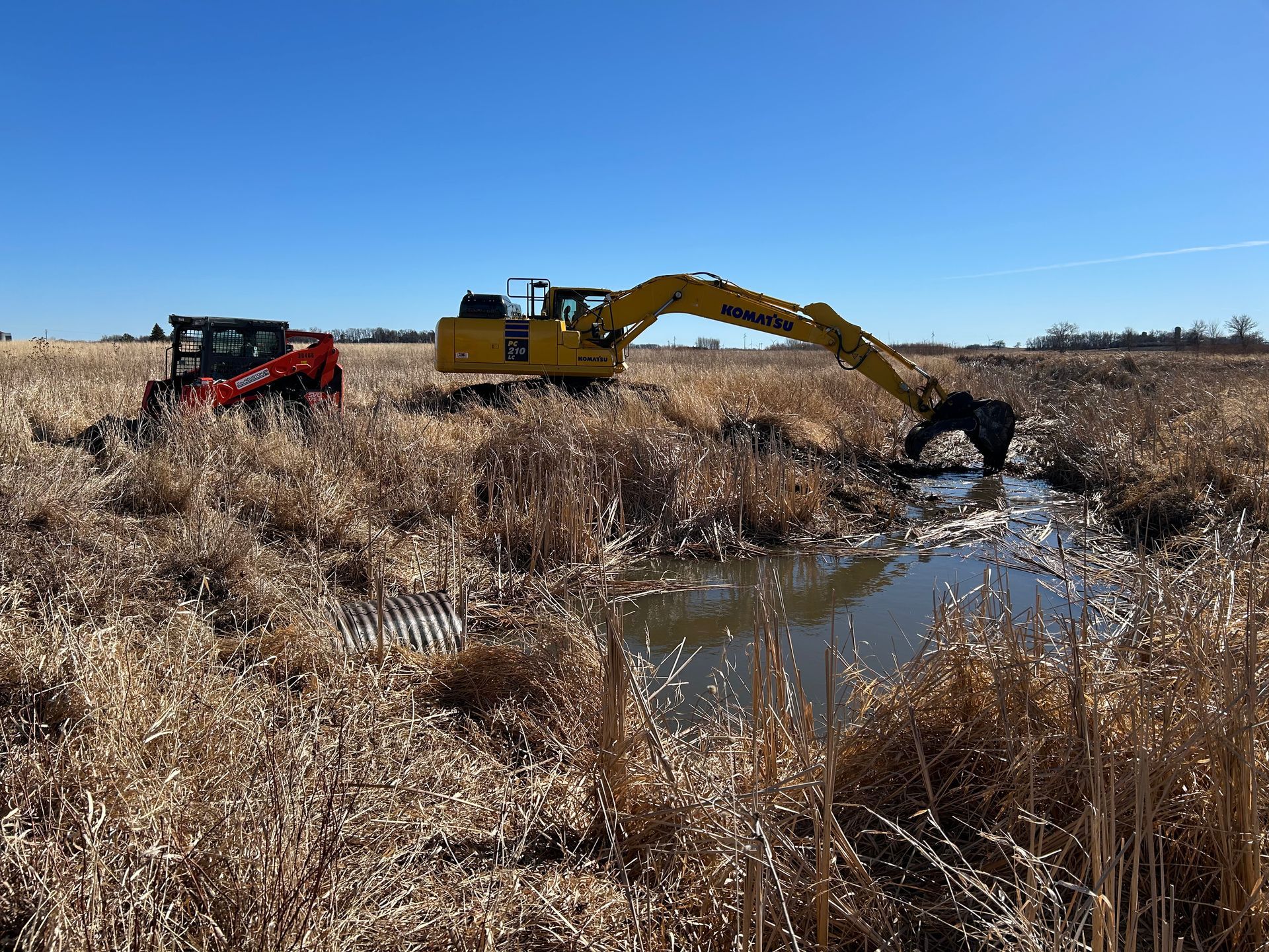 Two construction vehicles clearing a waterway in a field under a blue sky.