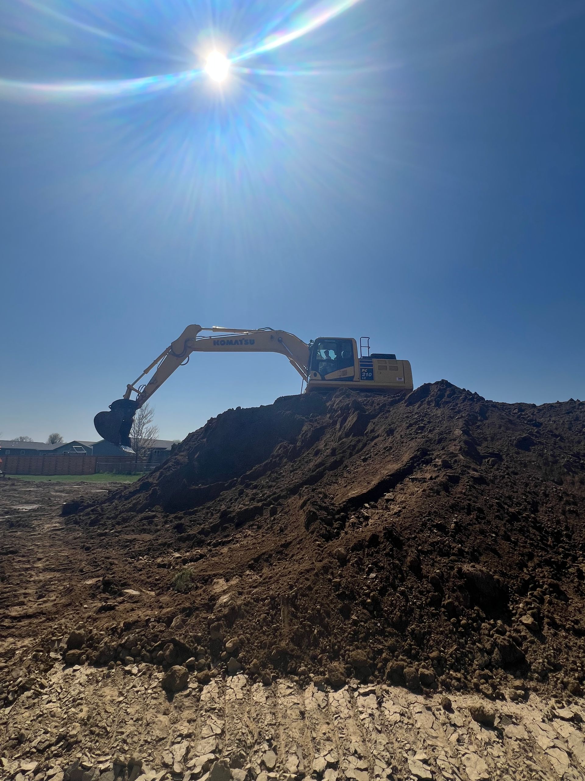 An excavator on a large dirt mound under a bright sun. Clear blue sky.