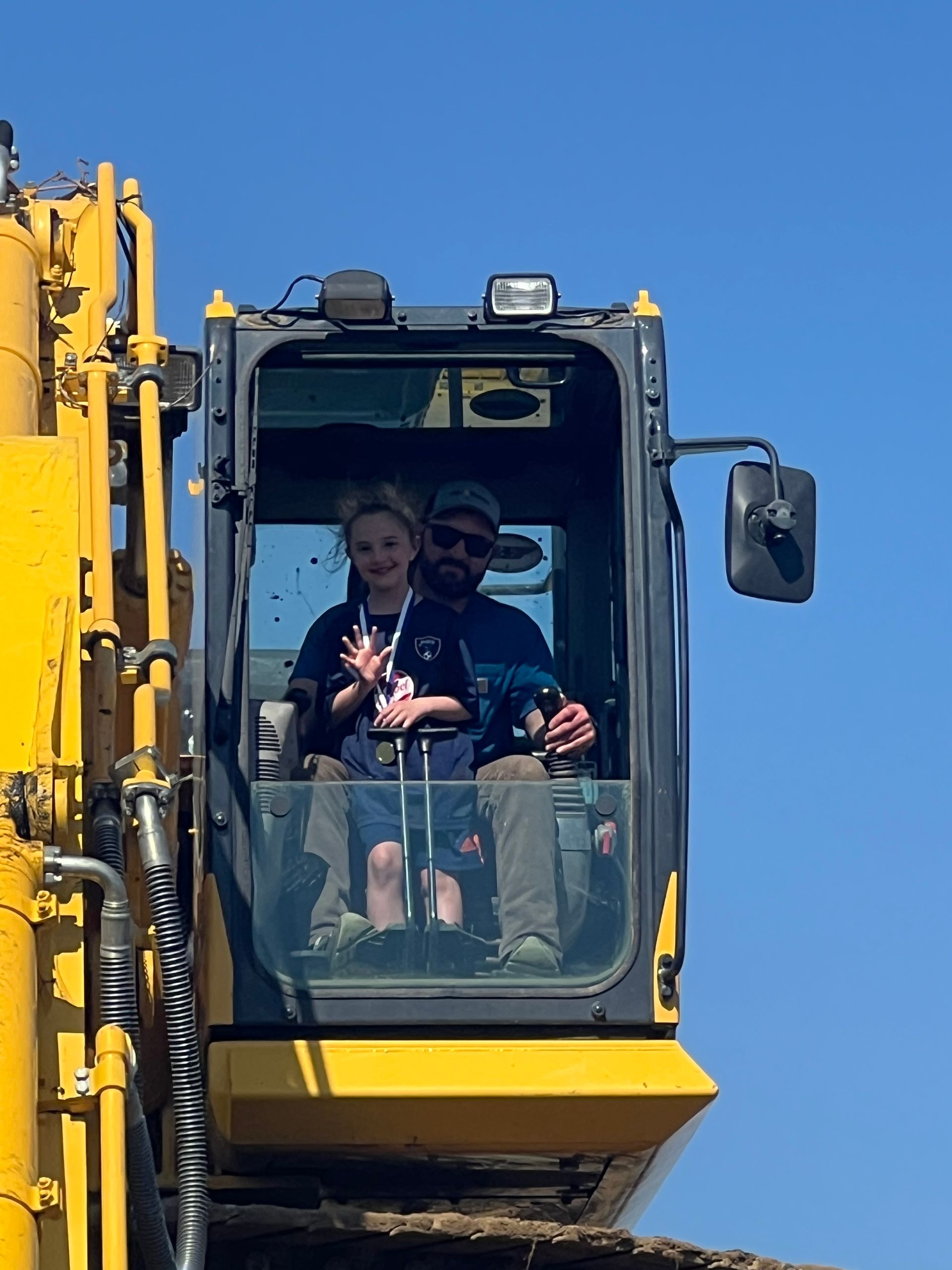 Man and child in yellow excavator cab, smiling against blue sky.