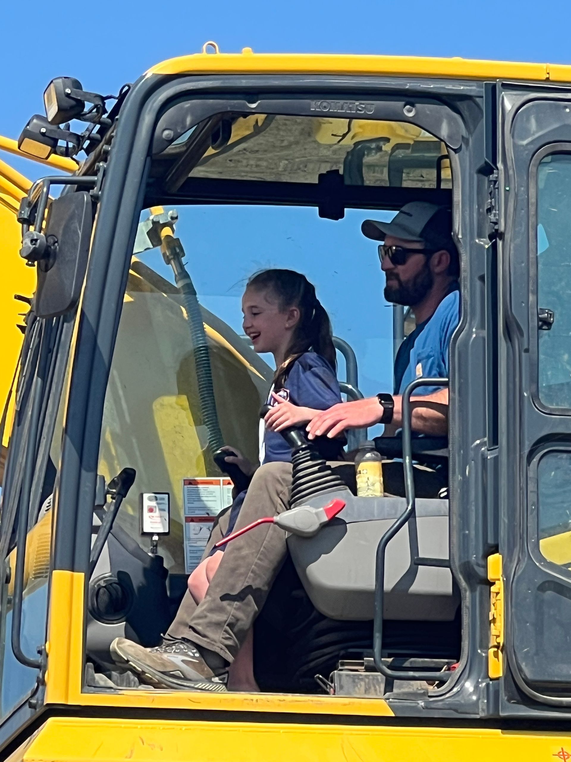 Man and girl in the cab of a yellow excavator; girl is smiling, man is operating the controls.