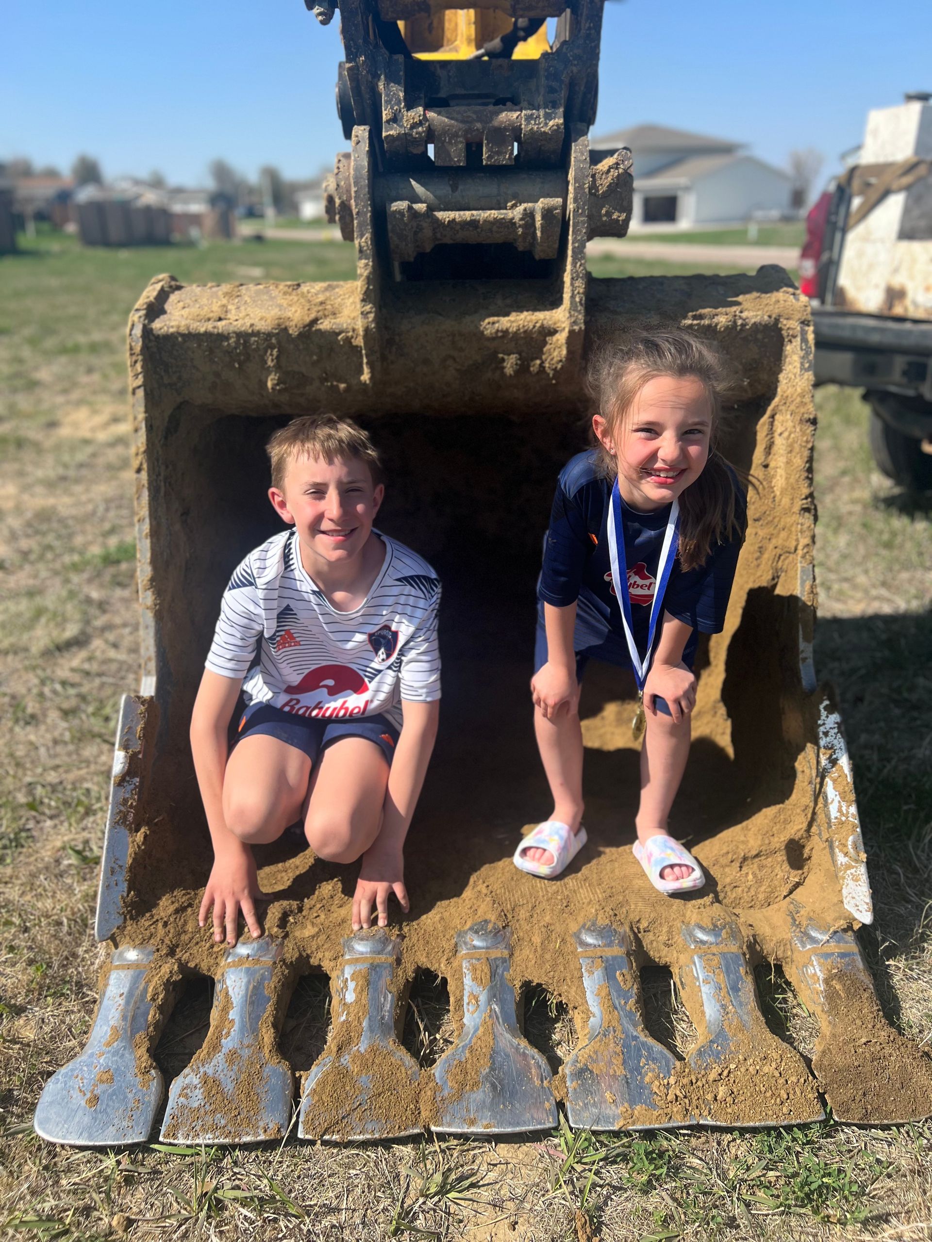 Two children inside excavator bucket, smiling, in a sunny outdoor setting.