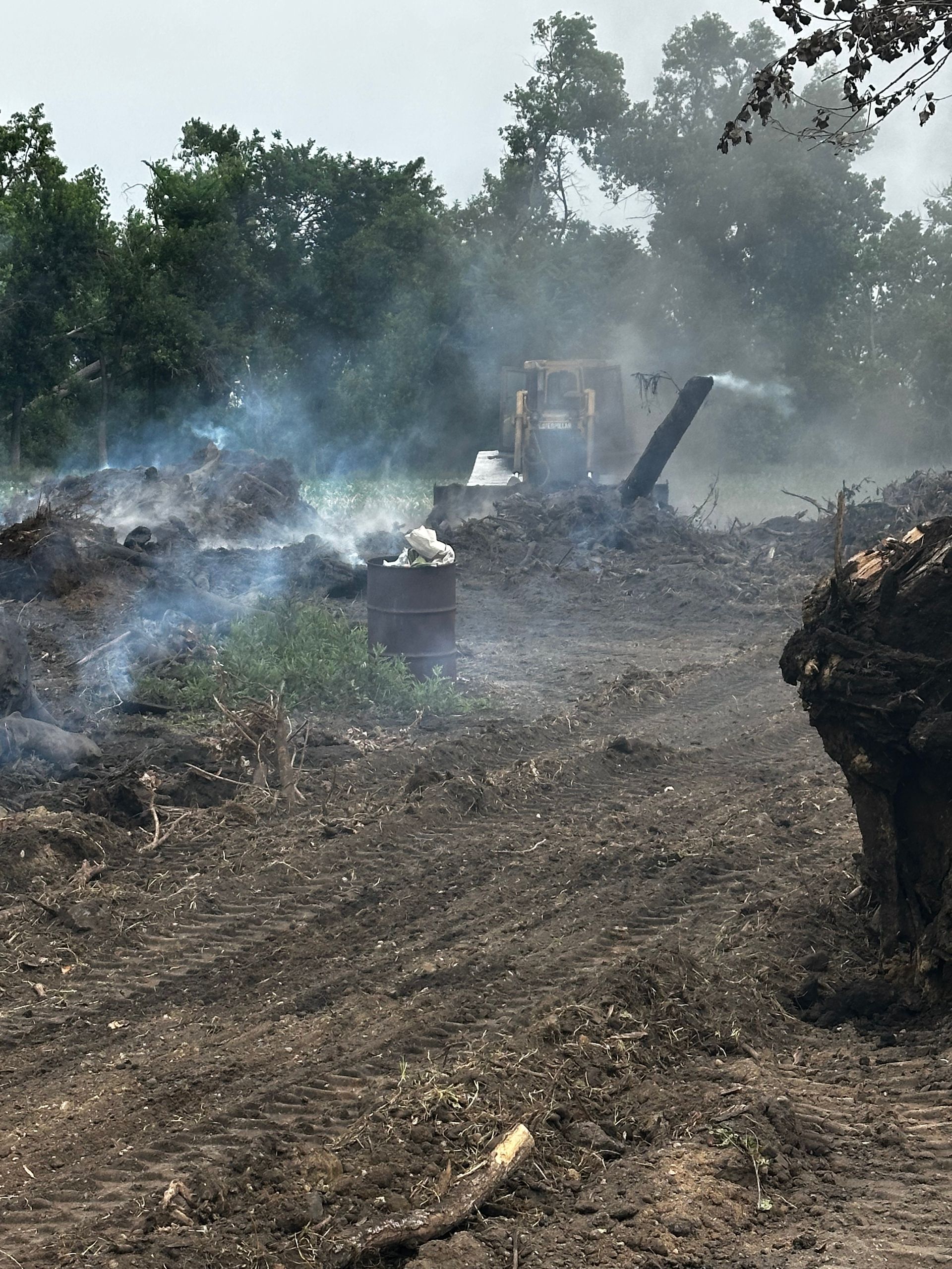 Smoldering debris pile with smoke, wood chipper in the background, surrounded by dirt and trees.