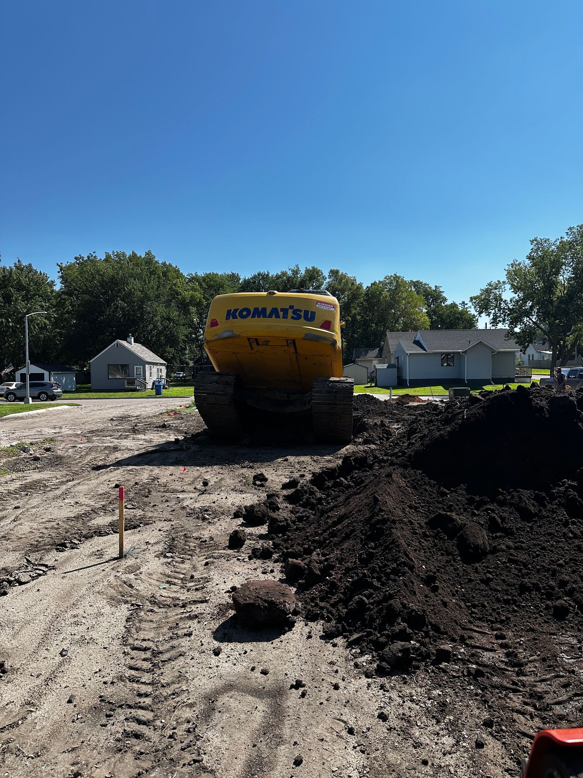 Yellow Komatsu excavator on a construction site; blue sky, dirt, and houses in the background.