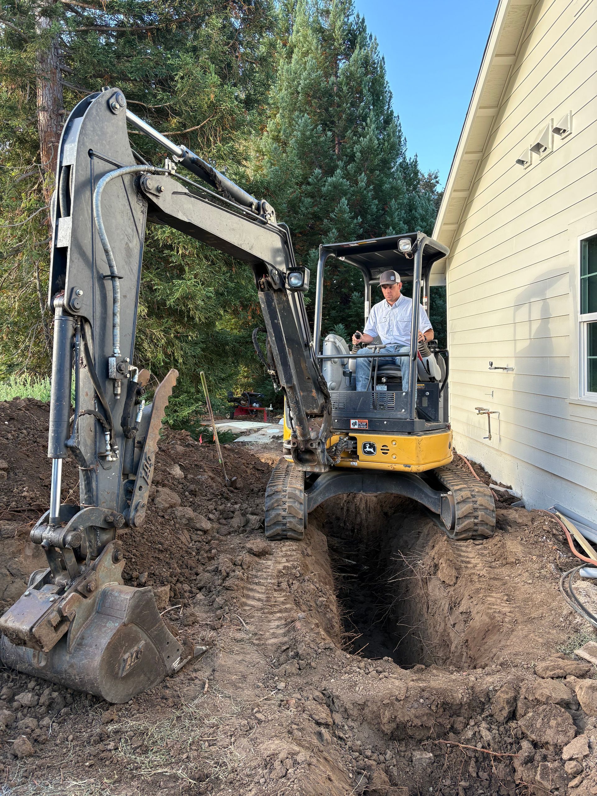 A construction worker operates a yellow mini-excavator to dig a trench alongside a beige house.
