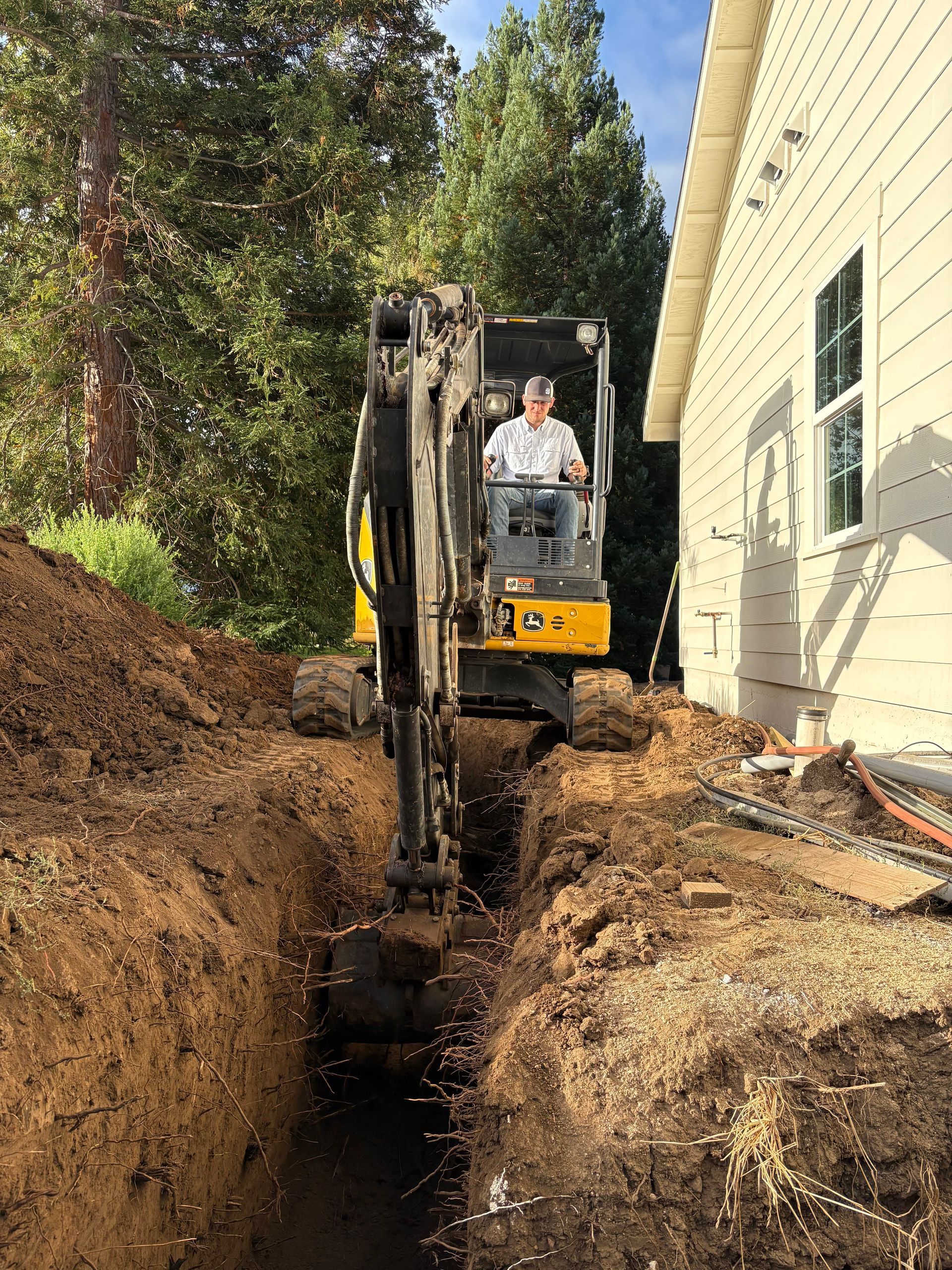 A person operates a yellow mini excavator to dig a trench along the side of a house near a wooded area.