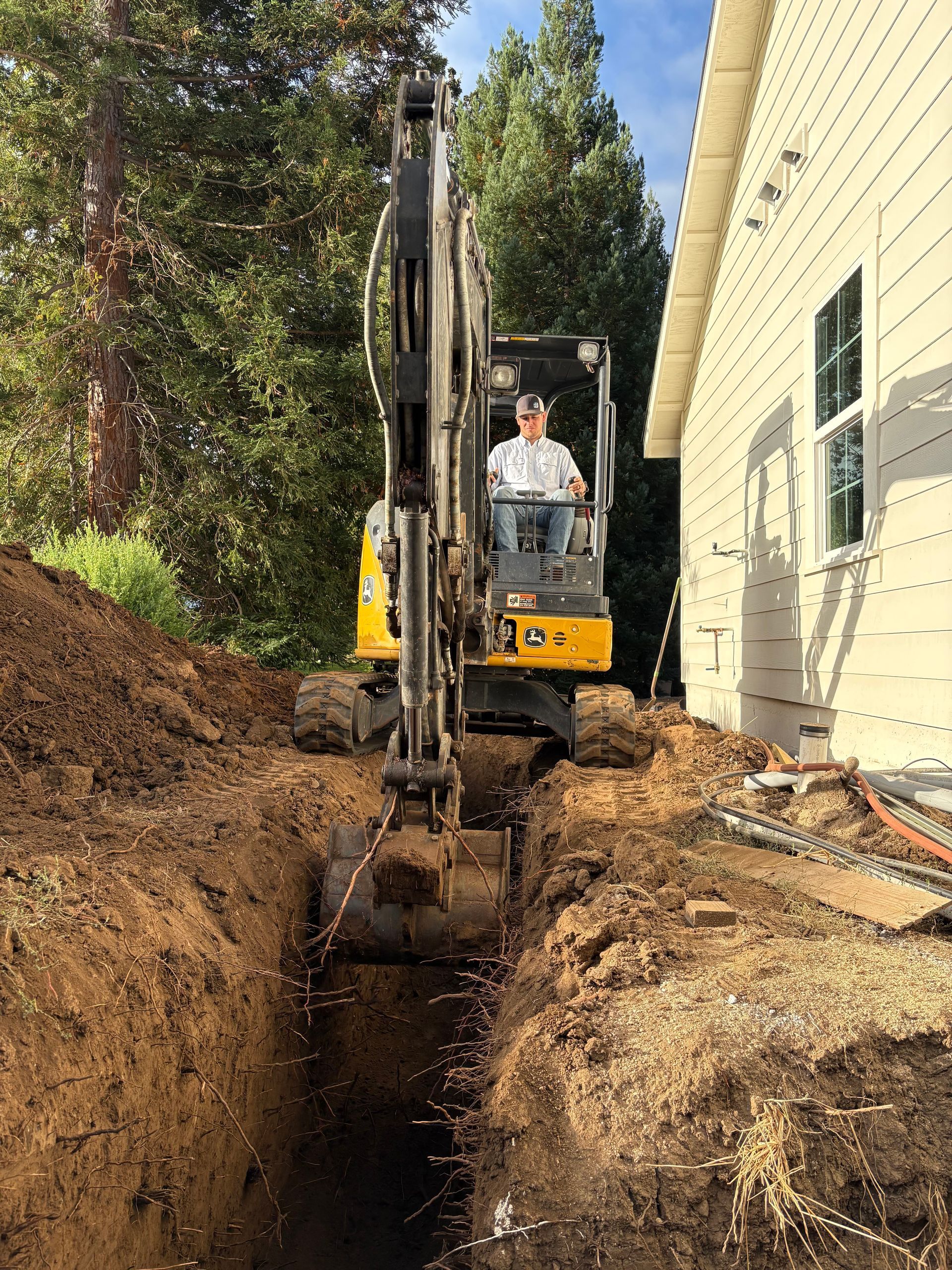 A person operates a yellow excavator digging a narrow trench beside the exterior wall of a house.