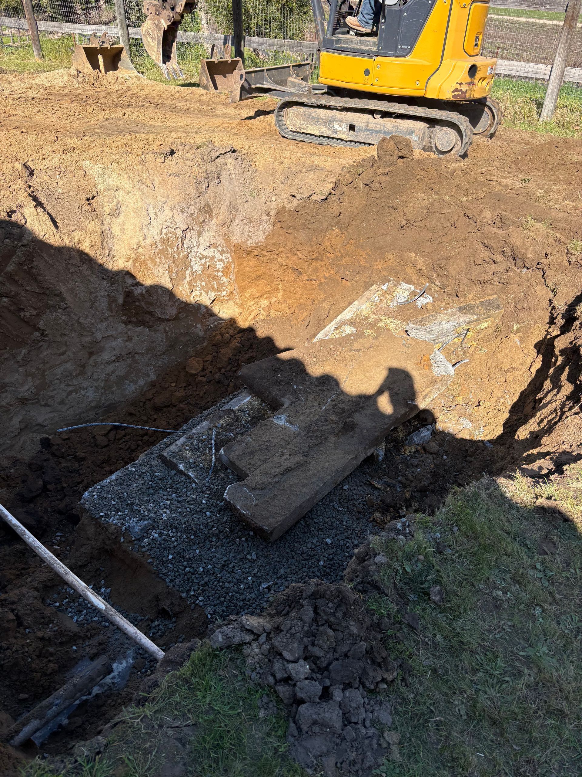 A yellow excavator is parked near an excavated trench in a dirt field, revealing a concrete structure underground.