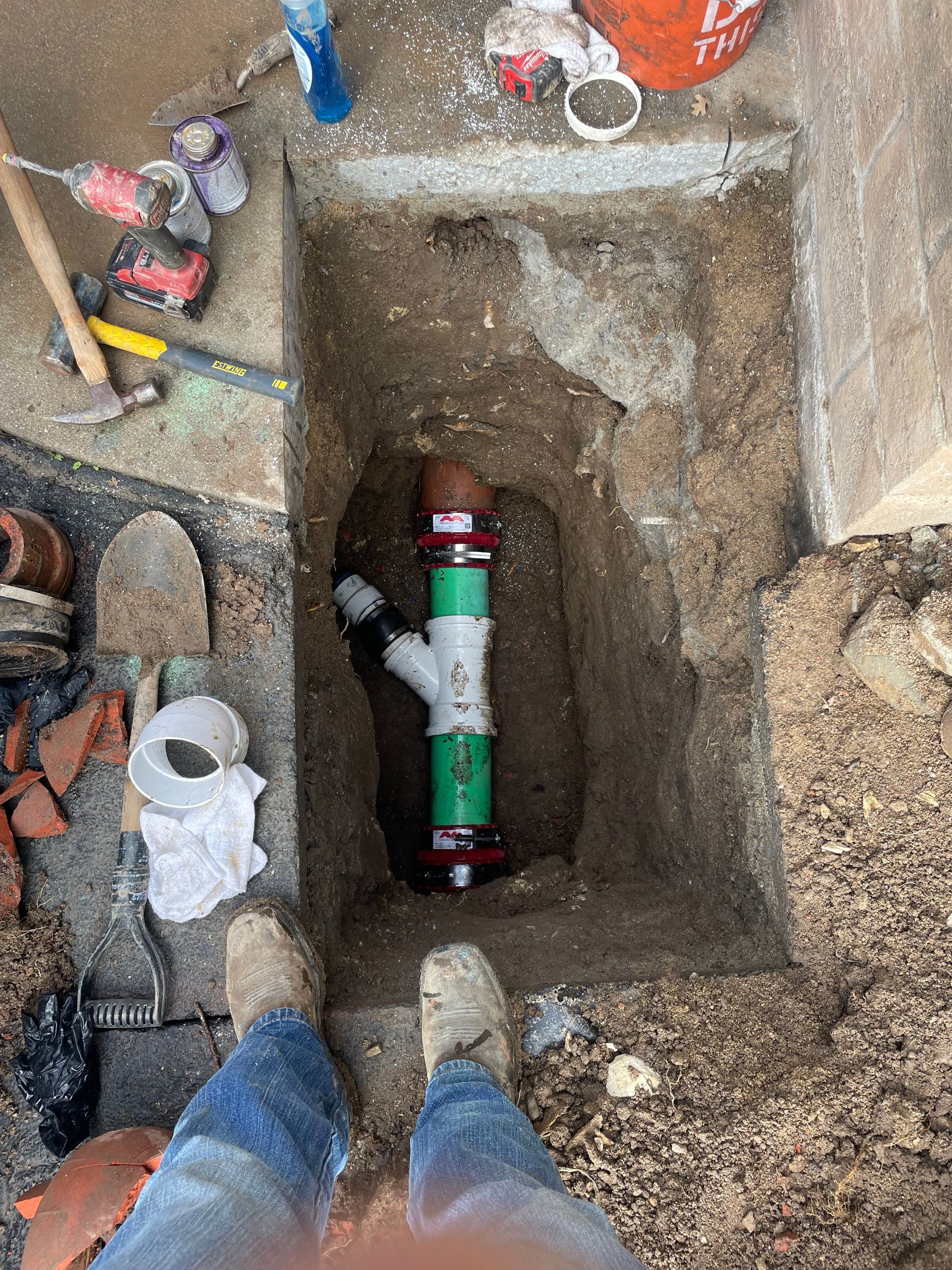 A person stands over a trench containing a newly installed green and white pipe Y-fitting connected to existing piping.