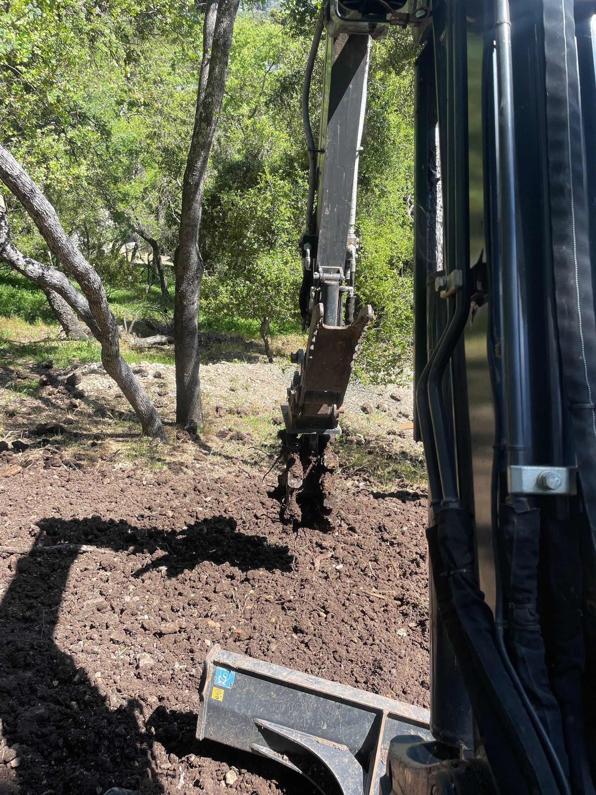 An excavator arm and claw attachment operate over tilled, dark soil in a wooded area.