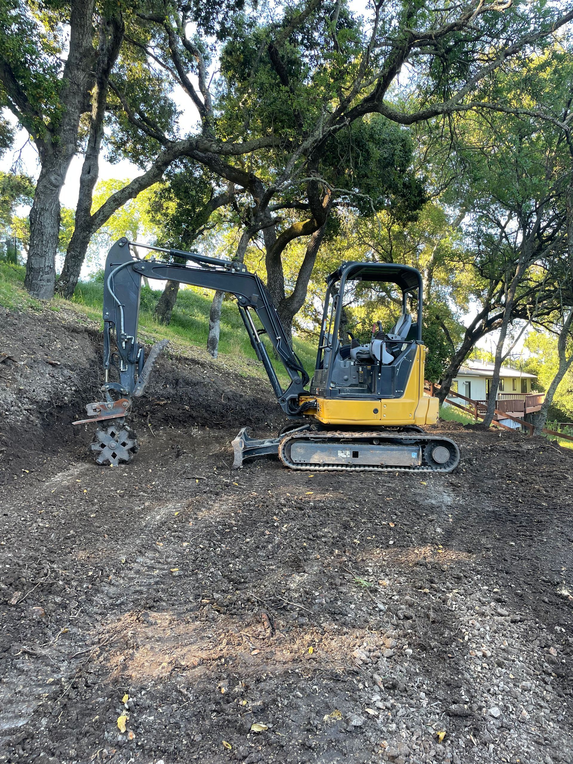 A yellow mini excavator sits on a dirt hillside under trees, with a mechanical attachment lowered toward the ground.