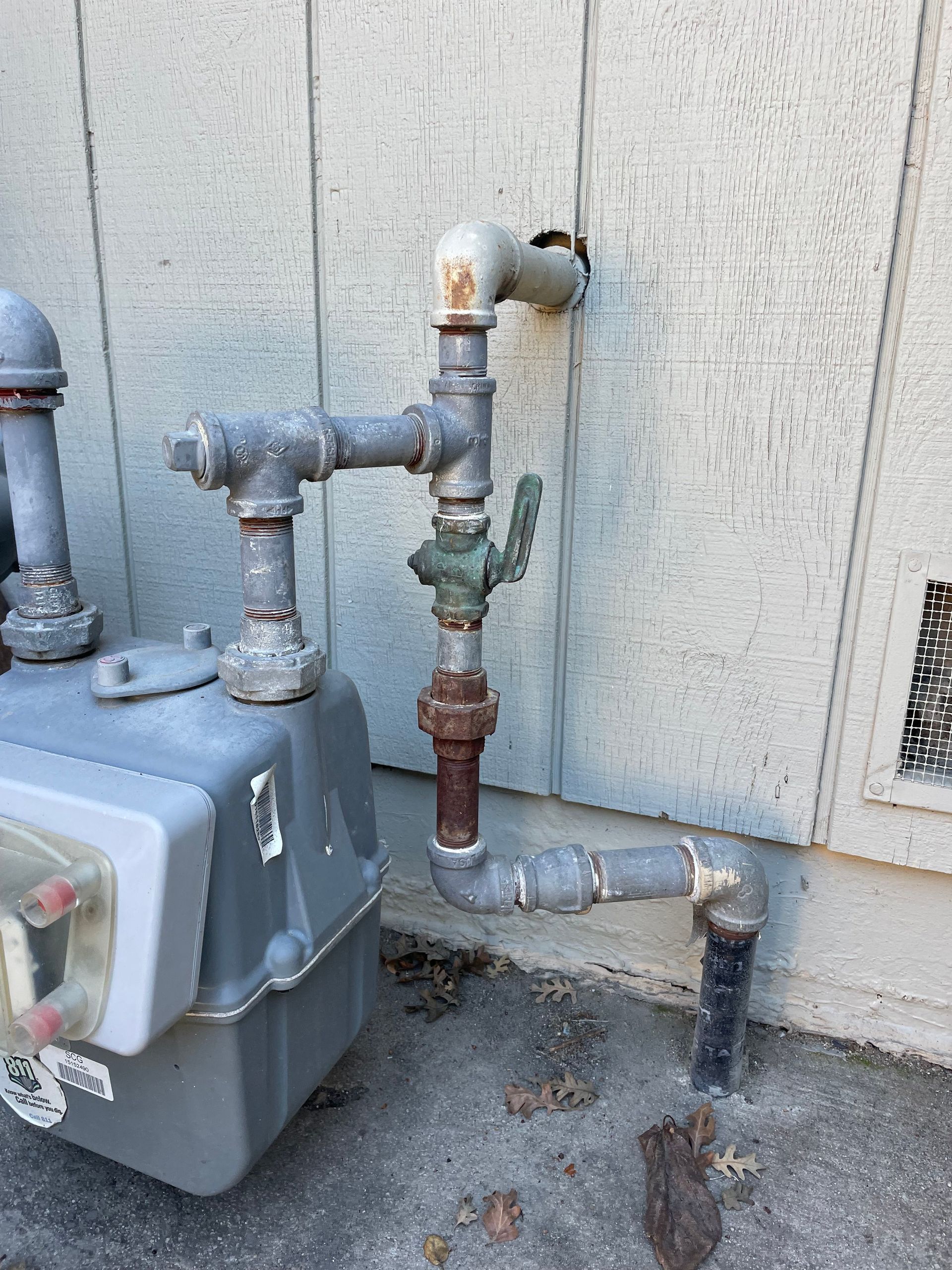 A grey natural gas meter and pipe assembly connected to the side of a building with light-colored wooden siding.
