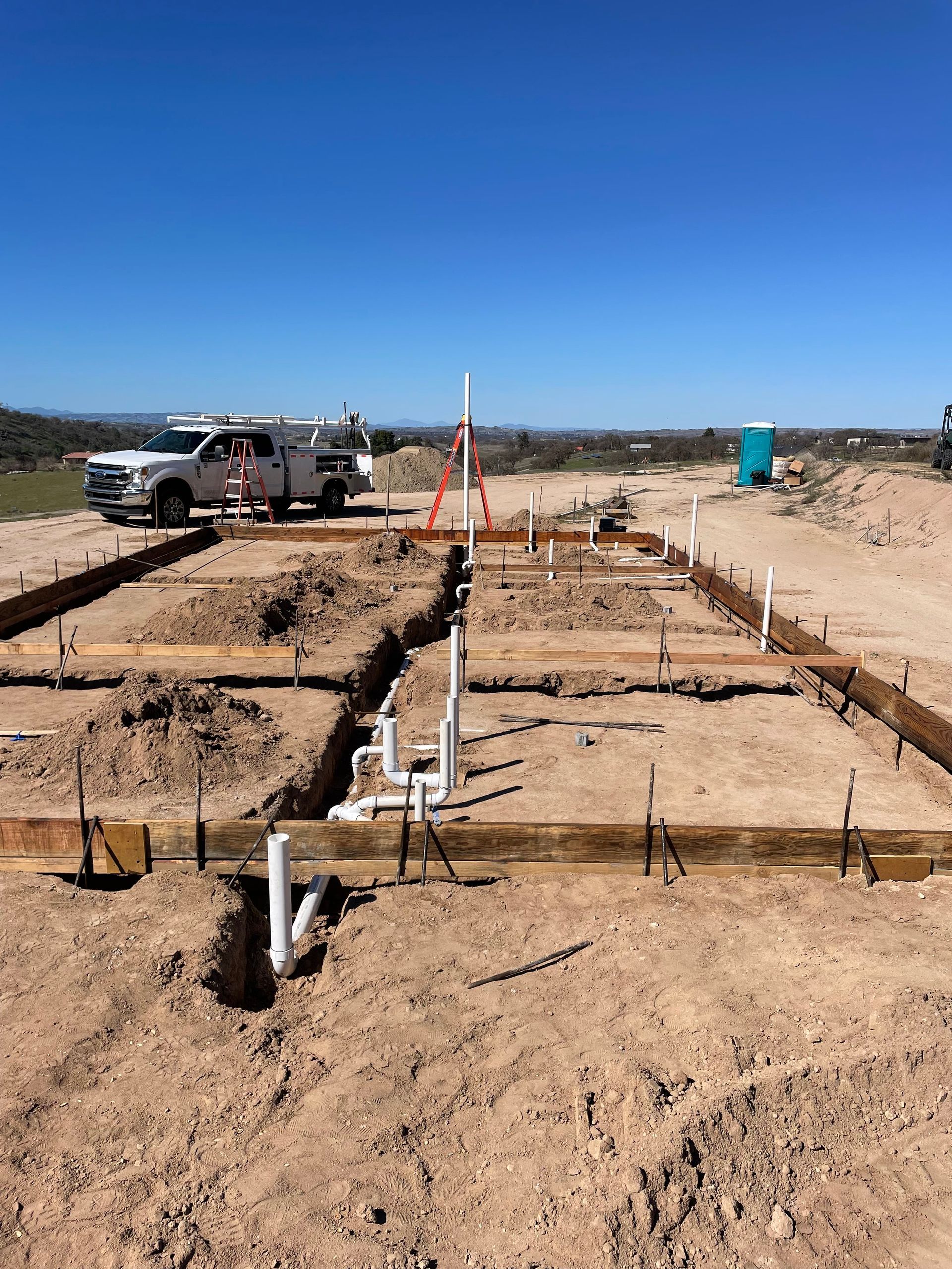 A construction site with foundation trenches, plumbing pipes, wooden form boards, and a utility truck under a blue sky.