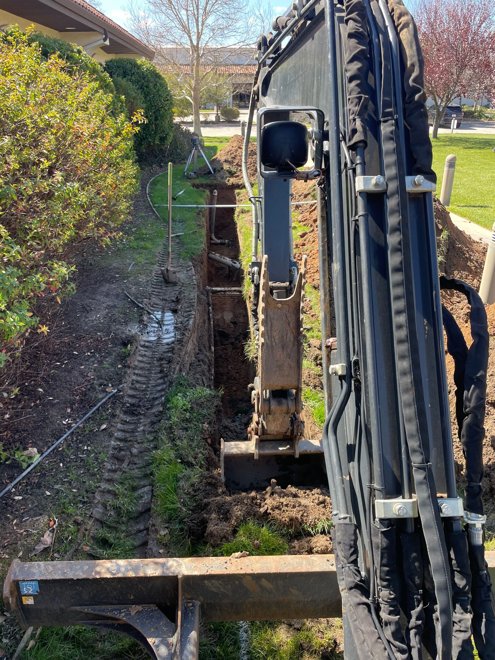 An excavator digs a long, narrow trench in a landscaped area beside a building.