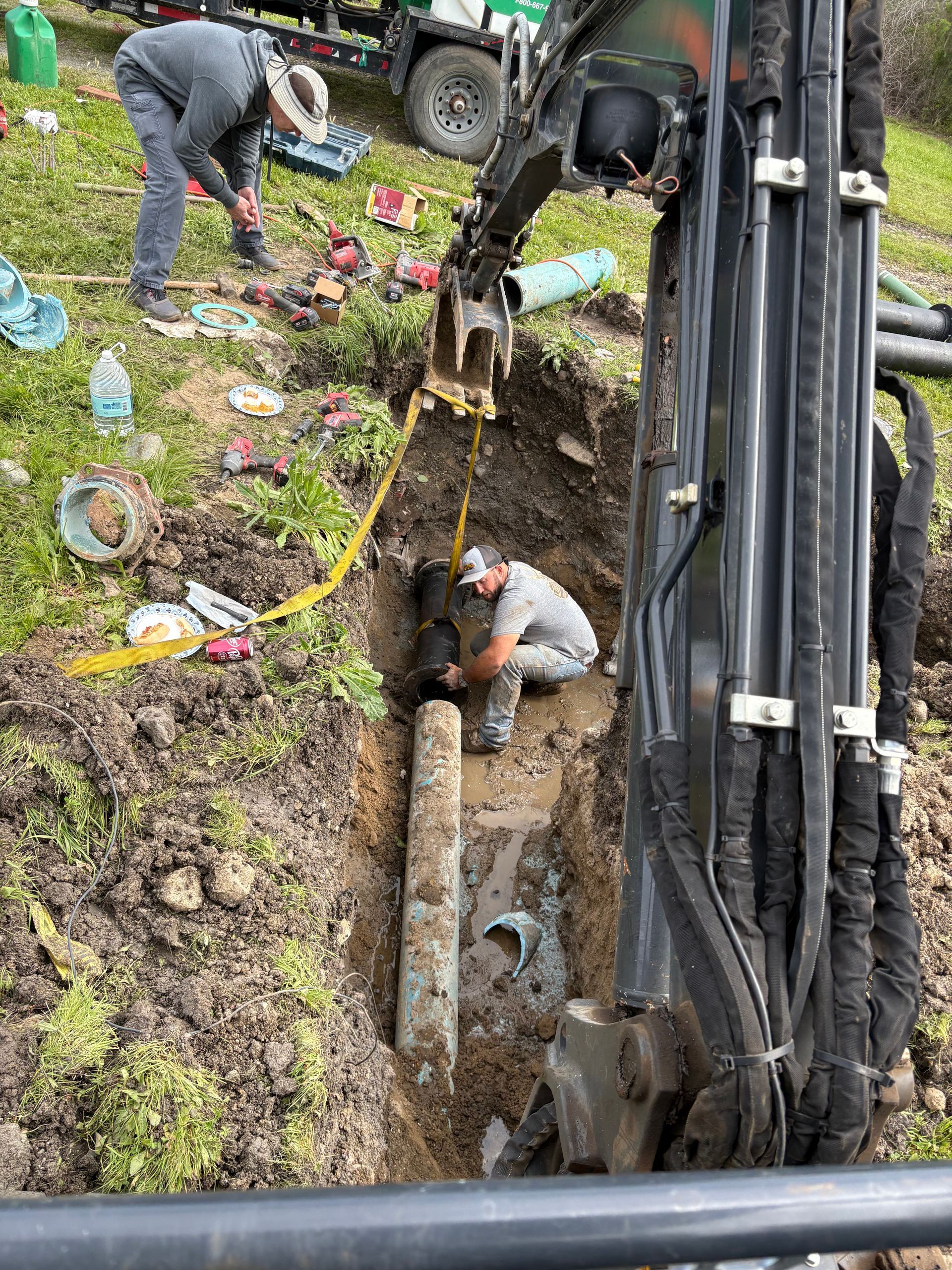 A construction worker crouches in a trench next to an excavator to repair an exposed underground pipe outdoors.