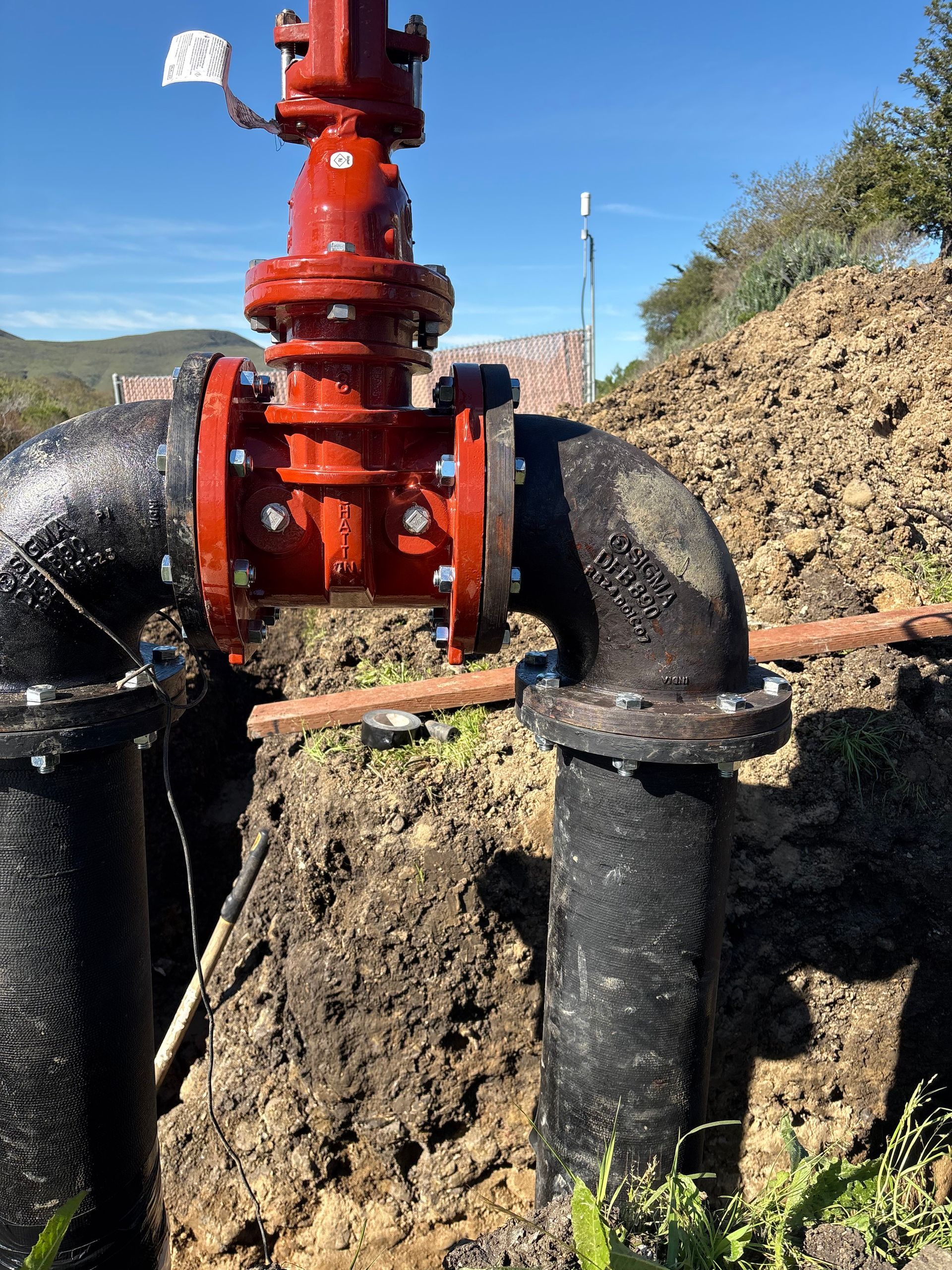 A bright red gate valve connected to two black industrial pipes in an outdoor dirt trench.
