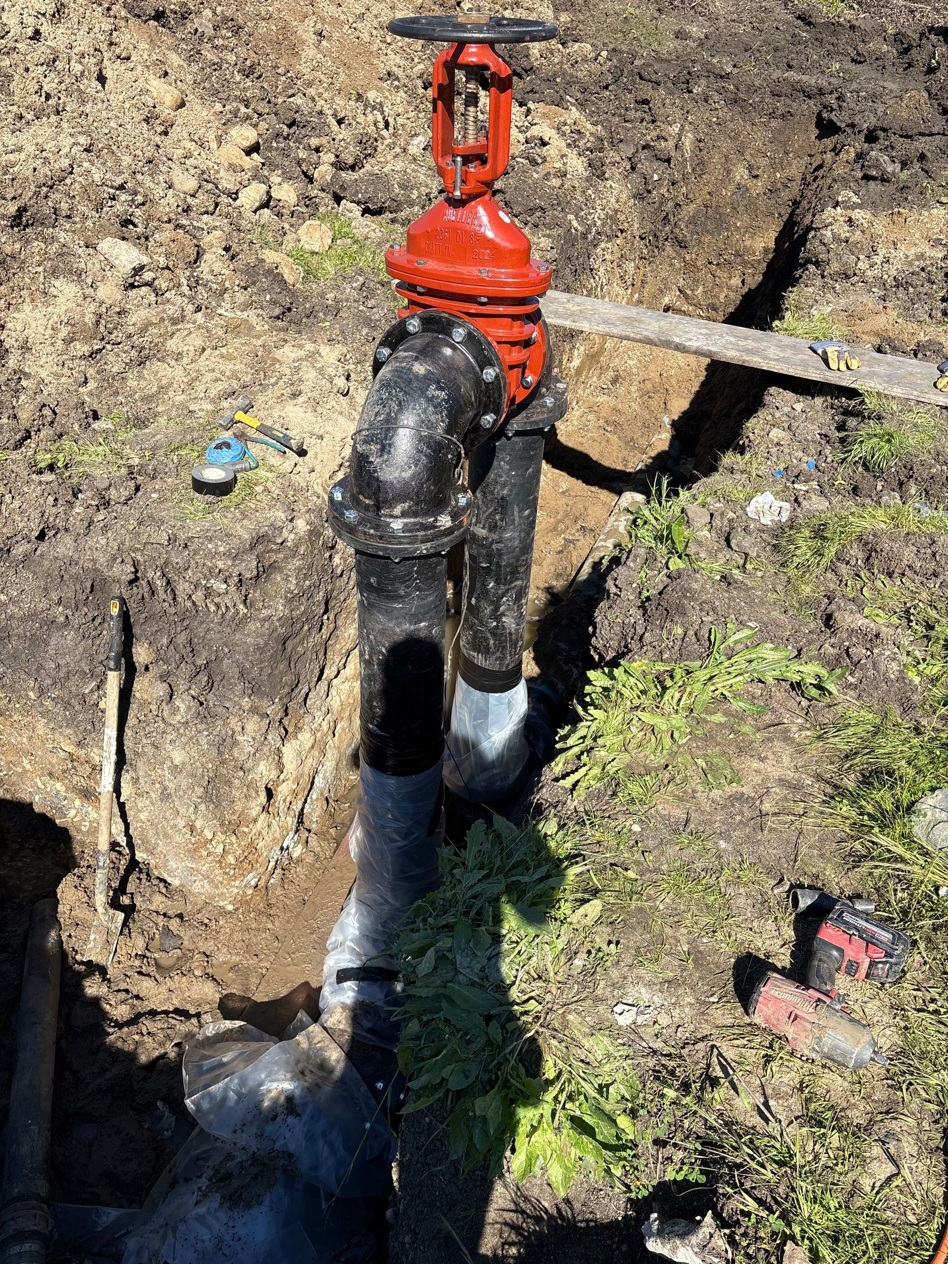 A red water valve installed on black pipes in a trench with construction tools nearby in an outdoor area.
