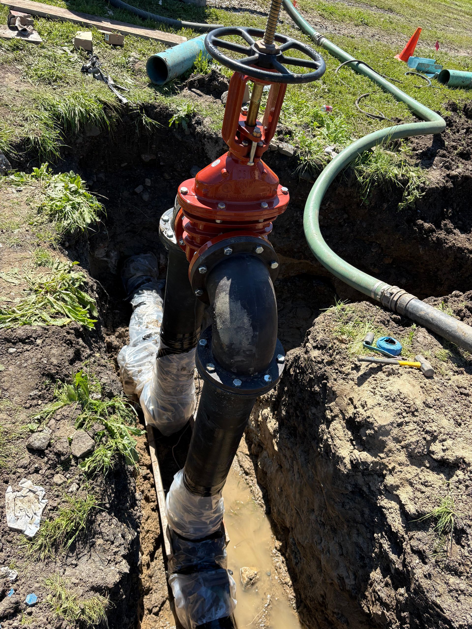 A bright red metal valve atop a black pipe assembly installed in an open muddy trench outdoors.