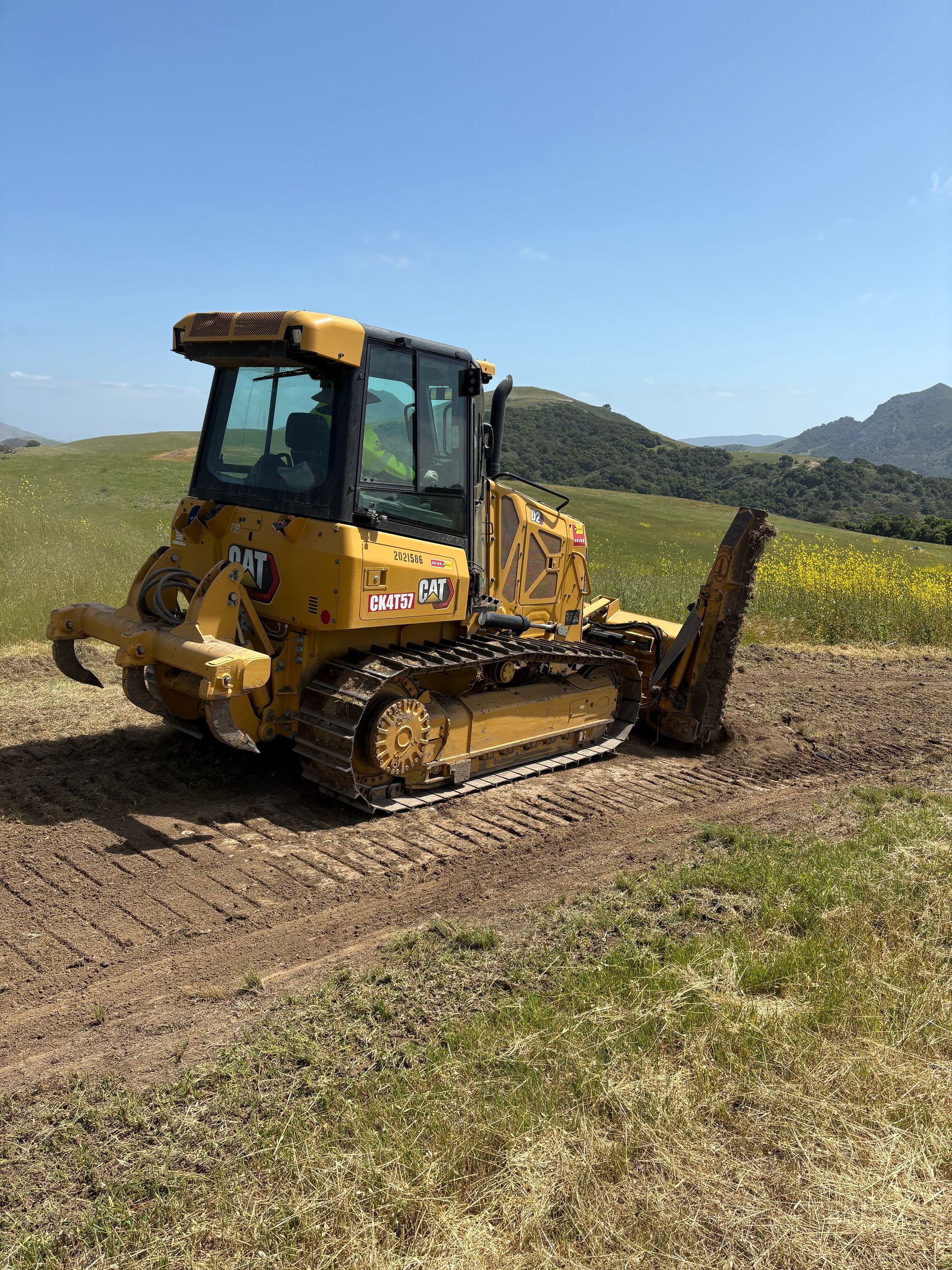 A yellow Caterpillar bulldozer on a dirt path in a grassy, hilly field under a clear blue sky.