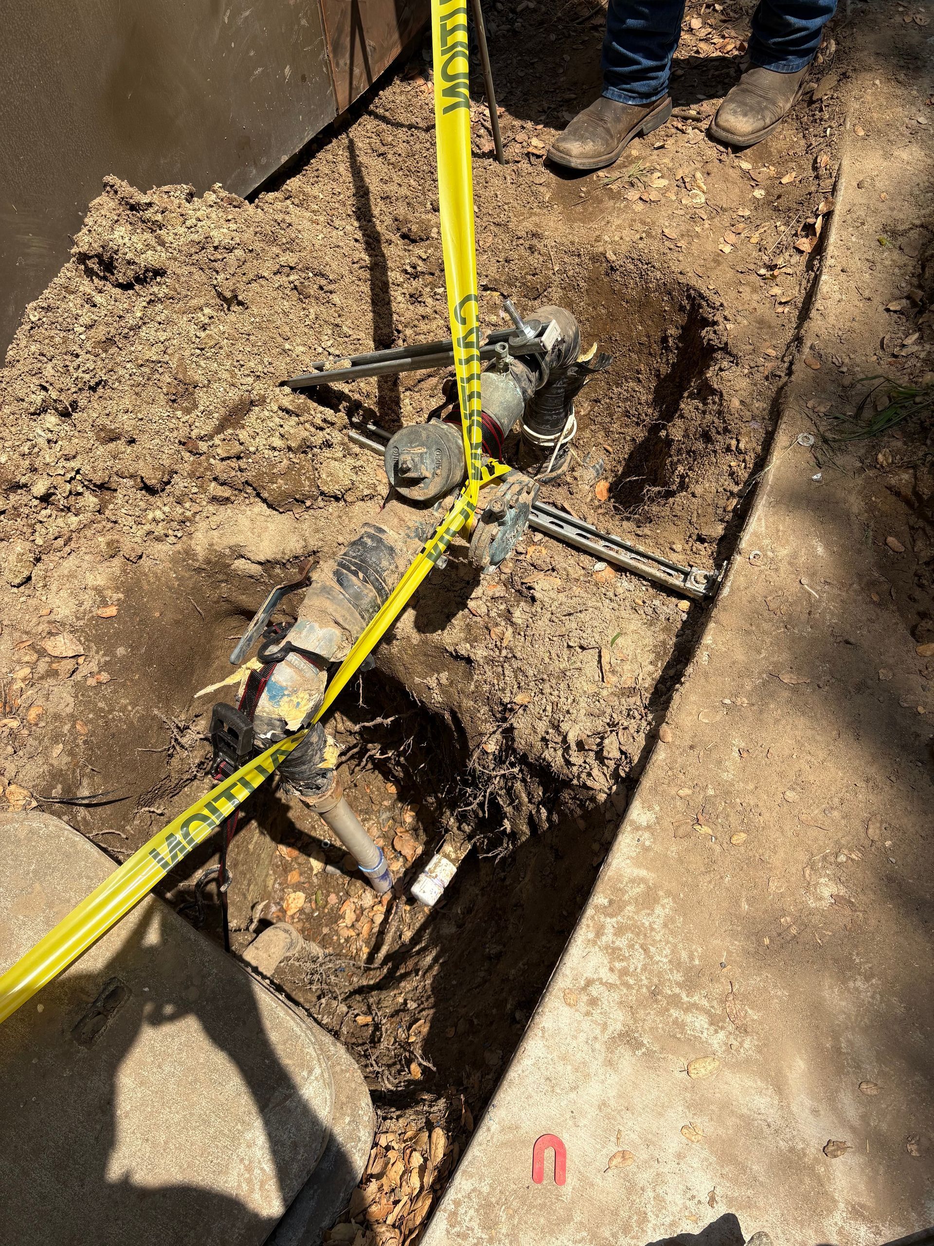 A worker's feet stand near an excavated water line with exposed valves and pipes, marked by yellow caution tape.